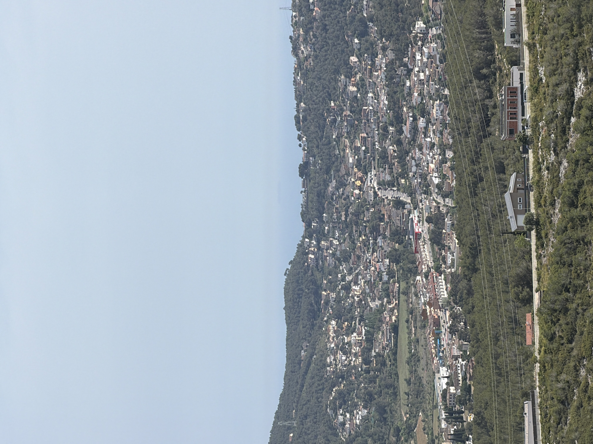 This panoramic photograph, captured in Olèrdola, Spain, showcases a vast residential landscape built into and around wooded hills under a clear, bright sky. The upper third of the image is dominated by a pale blue sky, indicating serene, sunny weather, likely midday or early afternoon.
Below the sky, a distant, undulating ridge line, thickly covered in dark green trees, forms the horizon. As the land descends towards the foreground, the hillsides become more extensively populated. Numerous houses, featuring light-colored facades and reddish-brown tiled roofs, are densely clustered, forming a sprawling town or village that blends with the natural contours of the terrain. The vegetation primarily consists of dense, evergreen trees, characteristic of a Mediterranean climate.
Further into the foreground, a prominent band of forest stretches horizontally, acting as a natural divider. Across this forested section, several parallel power lines are visible. Beyond this, to the right, a series of more contemporary buildings with distinct architectural styles are sparsely arranged along what appears to be a road or path, with further dense vegetation on a steeper rise beyond them. The overall scene depicts a harmonious, yet extensive, integration of urban development within a hilly, wooded environment. No human activity or visible text is present.