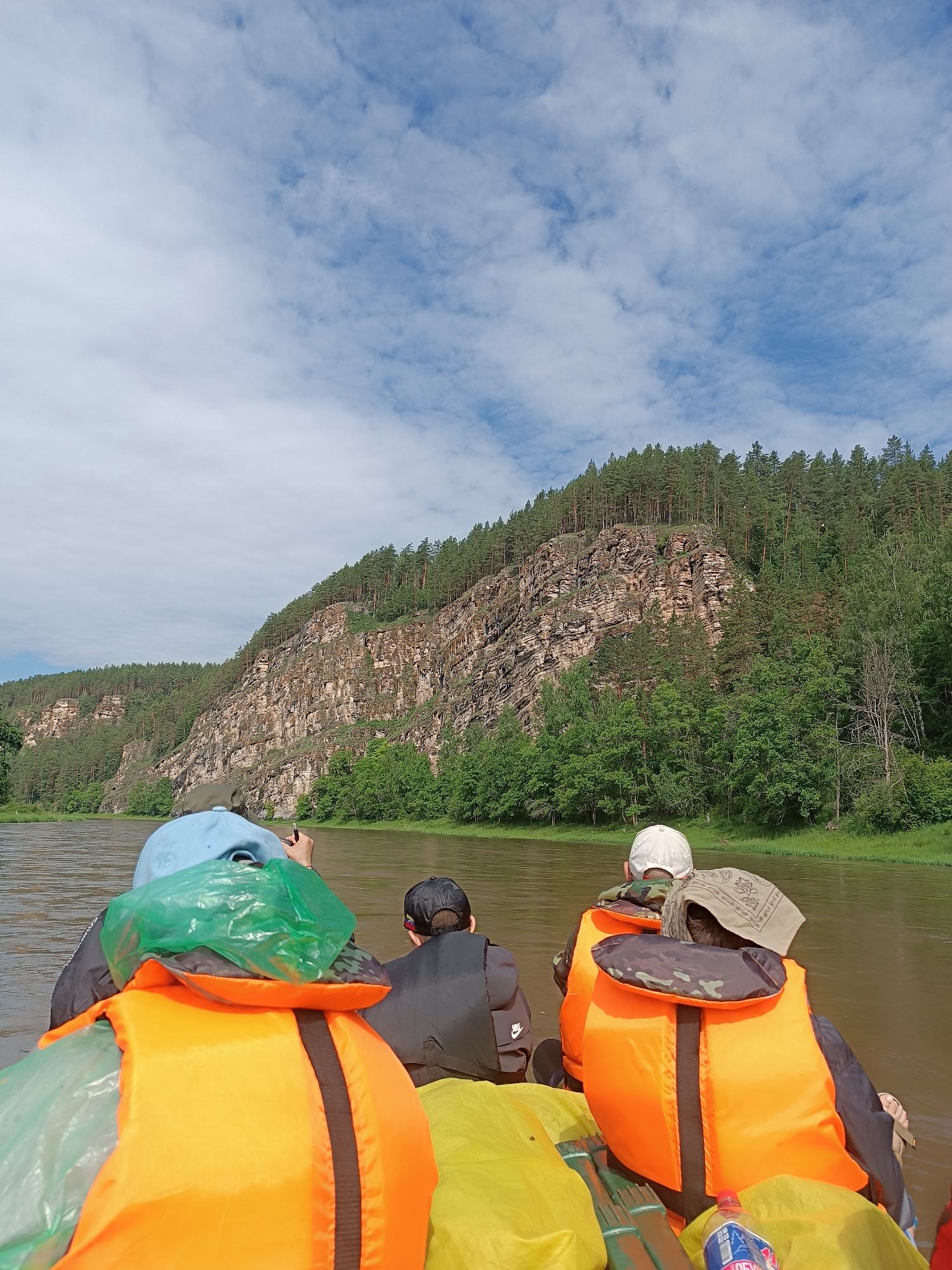 The image depicts a group of people on a raft or boat traveling down a brown river. The viewpoint is from behind the individuals, looking forward. The individuals are wearing life vests, with at least two in bright orange. One person wears a blue cap. The river is bordered by lush green trees and a steep, rocky cliff face covered in more trees. The sky is partly cloudy with patches of blue. The scene appears to be a natural landscape, likely a wilderness area or national park. No specific city landmarks are visible.