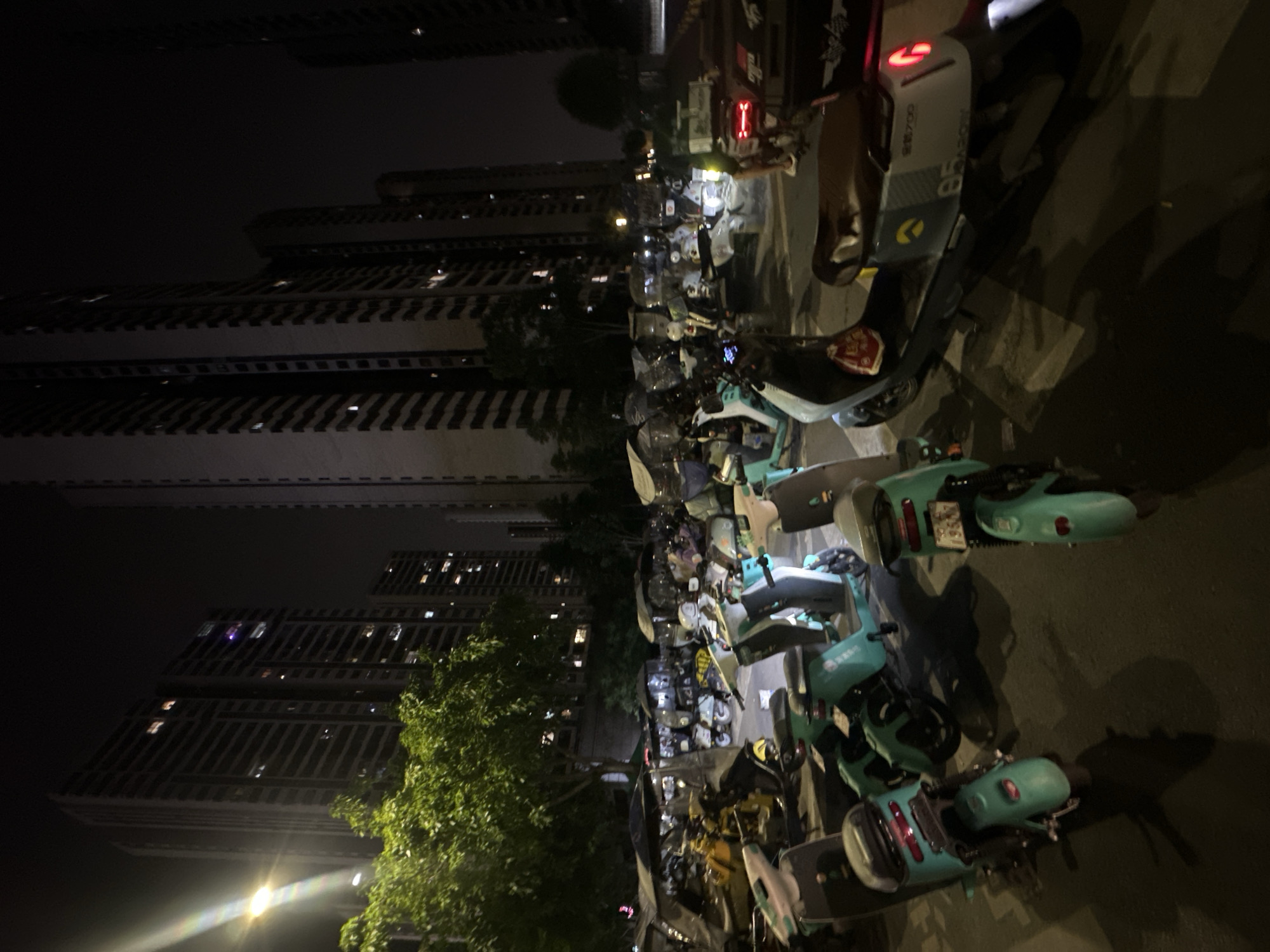 The image depicts a nighttime scene in Chongqing High-tech Zone, China. In the foreground, a large group of shared electric scooters are parked on the side of a road. The scooters are various colors including green, yellow and white. In the background, tall apartment buildings can be seen in the dark night sky. The lighting conditions are dim. Some trees are visible next to the buildings.