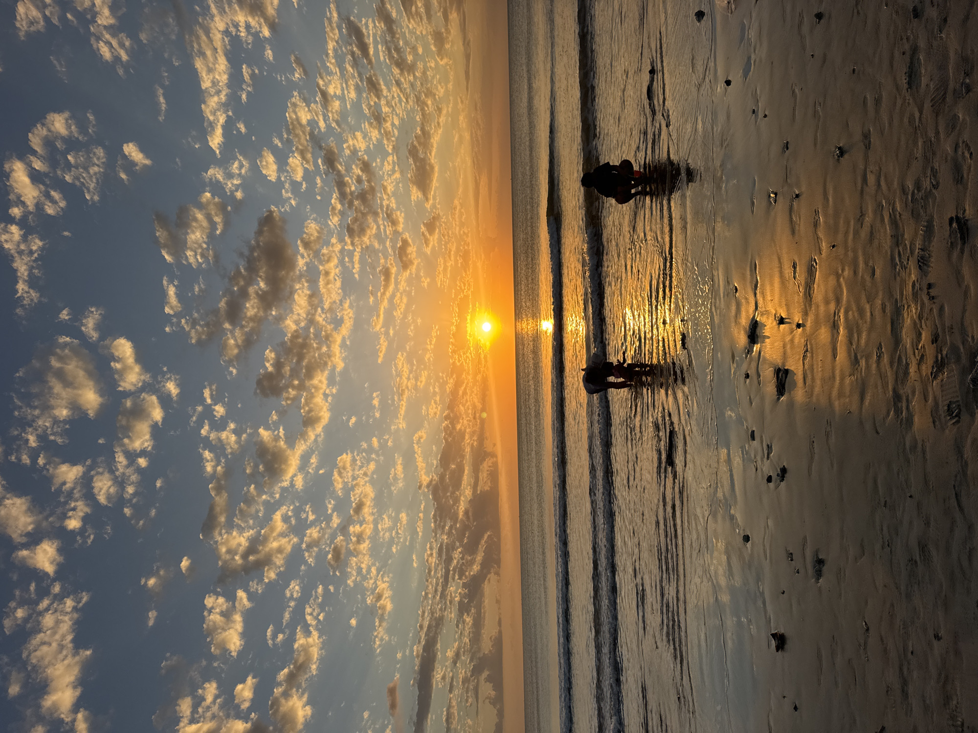 This image depicts a sunset scene on a beach in Kuta Selatan, Indonesia. The sun is a bright orange orb on the horizon, casting a golden glow across the sky and water. The sky is filled with scattered clouds that are illuminated by the setting sun. Gentle waves are rolling onto the shore, and the wet sand reflects the colors of the sunset. Two silhouetted figures are visible in the shallow water near the shore. One figure is crouched down, seemingly observing something in the water, while the other, slightly further away, is also crouched. The overall atmosphere is serene and picturesque.