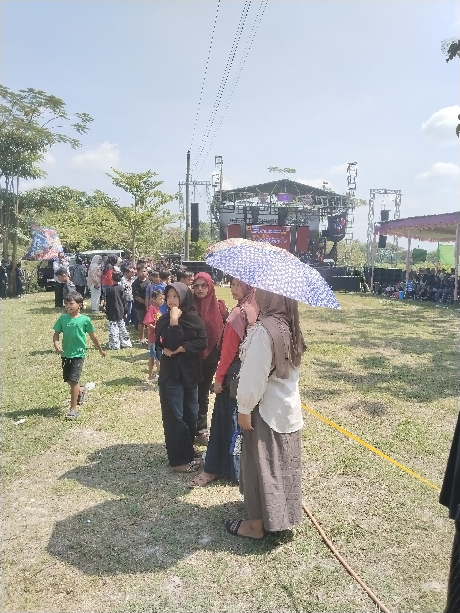 A crowd stands in a grassy field in Karanganom, Indonesia. A stage is in the background, with speakers and a canopy. Many people stand in a line; a person with an umbrella shields some of them. A boy in a green shirt walks towards the line. Trees line the perimeter of the field under a bright sky.