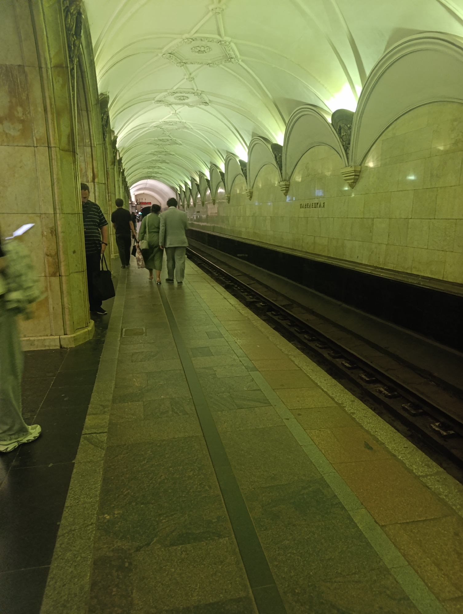 In a subway station, people stand on a platform beneath a white vaulted ceiling. The platform is lined with green stone tiles and has a set of train tracks running alongside it. A man in a striped shirt stands near a pillar, holding a black bag. Further down the platform, a couple in matching green outfits walks away from the camera. The walls of the station are covered in light-colored marble tiles. A sign with Russian lettering is visible on the wall. The overall lighting is dim and appears to be illuminated by artificial sources, casting a greenish hue on the scene.