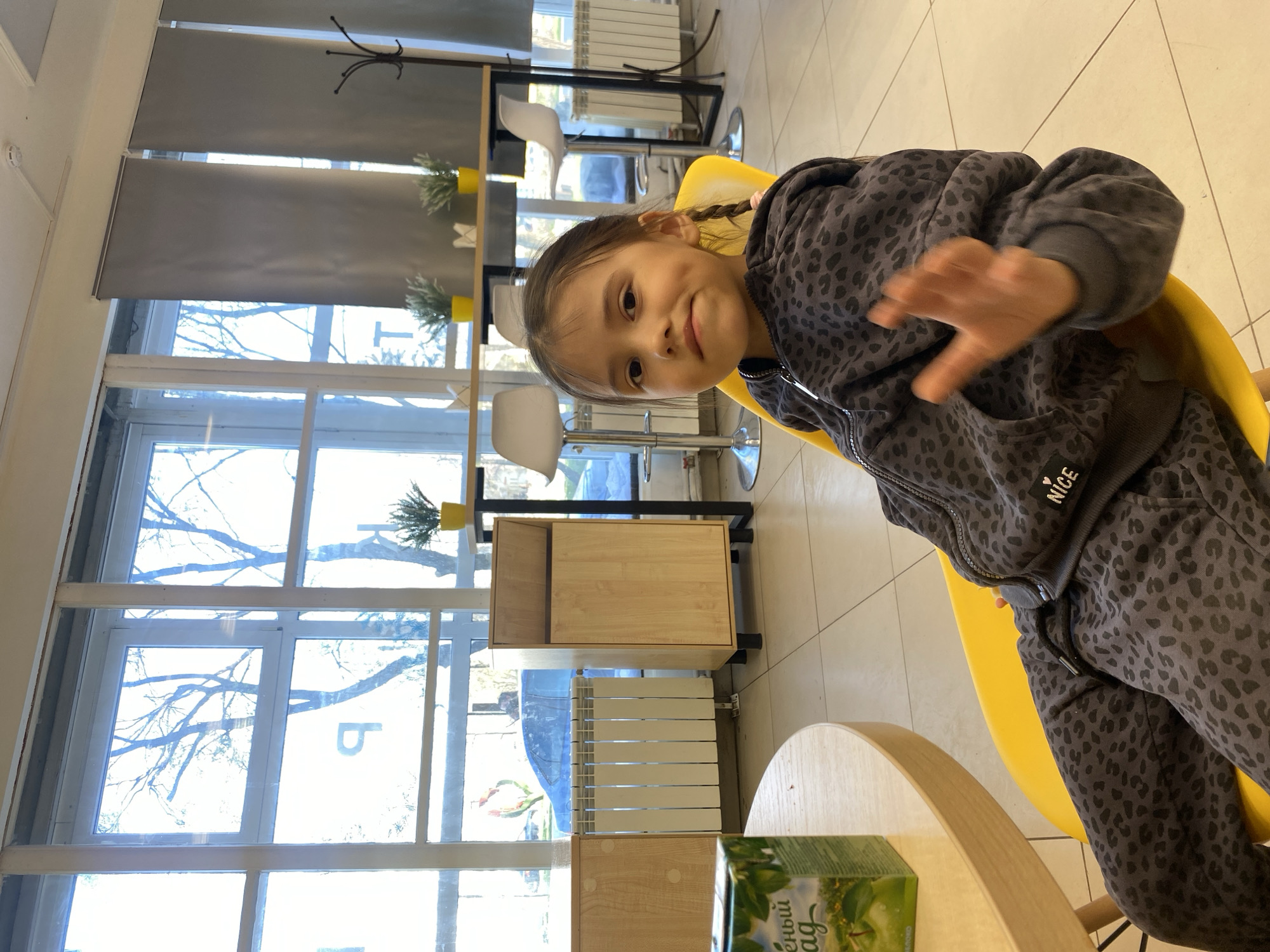 A girl sits in a yellow chair in a building with large windows. The girl is wearing a gray leopard-print tracksuit. She is smiling and waving. In the background, there are blinds, tables, chairs, a radiator, and a window with bare tree branches visible. The building is located in the city of Chaikovsky, Russia.