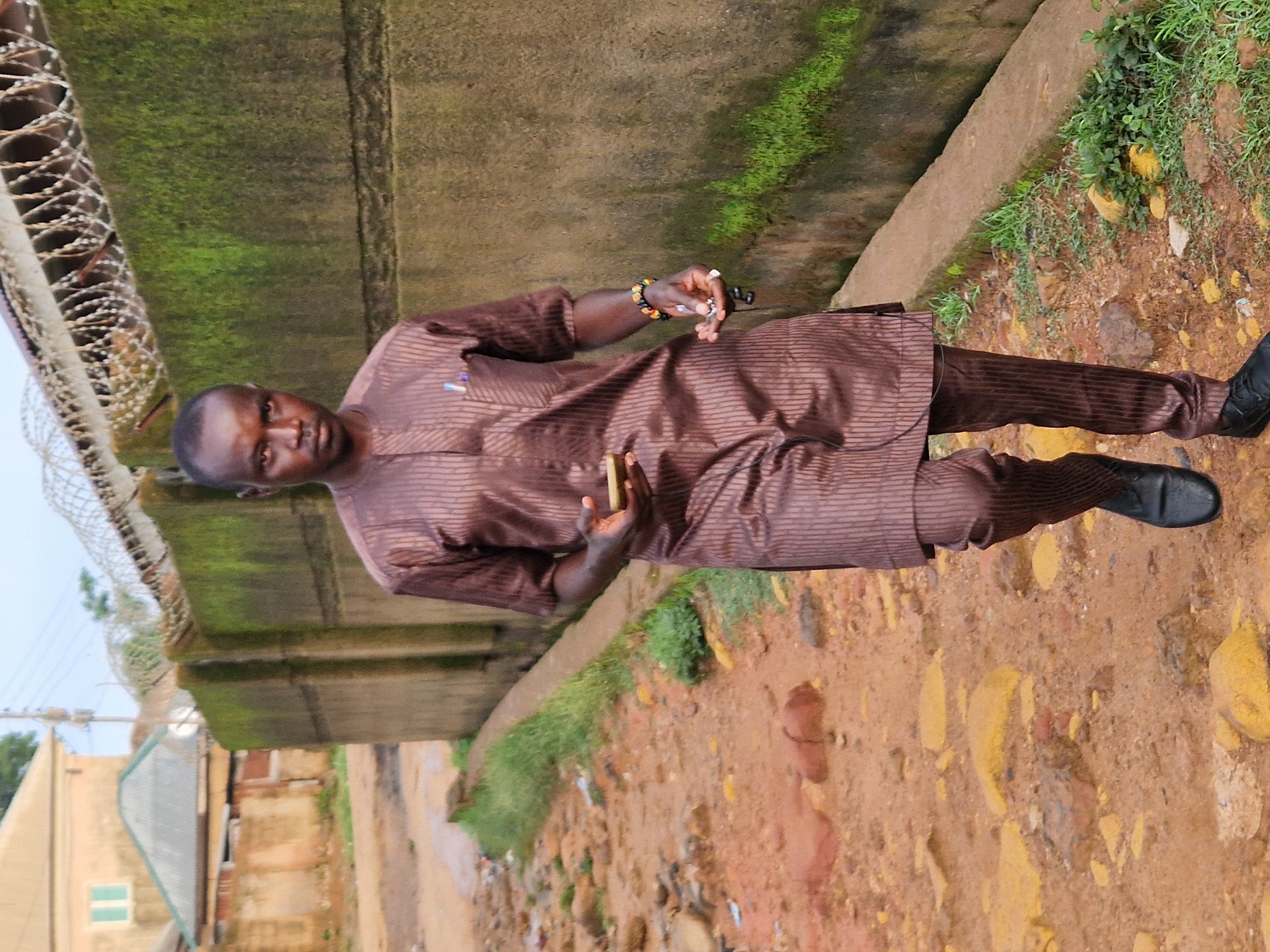 A full-body, eye-level, medium shot captures a man walking towards the camera on a dirt path lined with small rocks and sparse vegetation. The man, wearing a brown pinstriped traditional outfit, walks with his right hand holding a mobile phone and his left hand slightly extended, holding a small object. He has a bracelet on his left wrist. Behind him, a rough concrete wall covered in green moss is topped with barbed wire fencing. In the background, out-of-focus buildings and a utility pole are visible under an overcast sky. The scene appears to be in Kabong, Nigeria, characterized by its unpaved roads and rustic architecture.