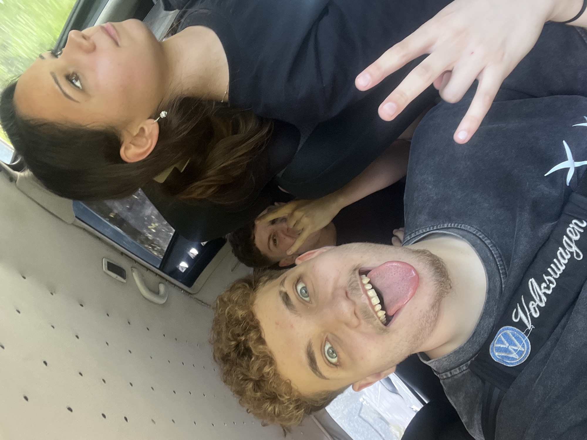 Three individuals are inside a vehicle. The person in the foreground, a male with curly blonde hair, is sticking his tongue out at the camera with his eyes wide open. He is wearing a dark grey t-shirt with a "Volkswagen" logo on a strap across his chest. To his left, partially obscured, is another male with short brown hair, making a peace sign with his fingers and a surprised expression. On the right, a female with dark hair pulled back in a ponytail looks up and to the side, wearing a dark t-shirt and a small earring. The interior of the vehicle appears to be a van, with a textured, cream-colored ceiling with small dark circular indentations. A white grab handle is visible near the window. The lighting suggests it is daytime, possibly overcast, with some light filtering in from the windows. The backdrop outside the windows is blurred, but appears to be greenery, suggesting a rural or suburban setting. There are no specific landmarks or indications that definitively place the location in Vlahovo, Bulgaria, but the overall scene is consistent with being in a vehicle in a community.