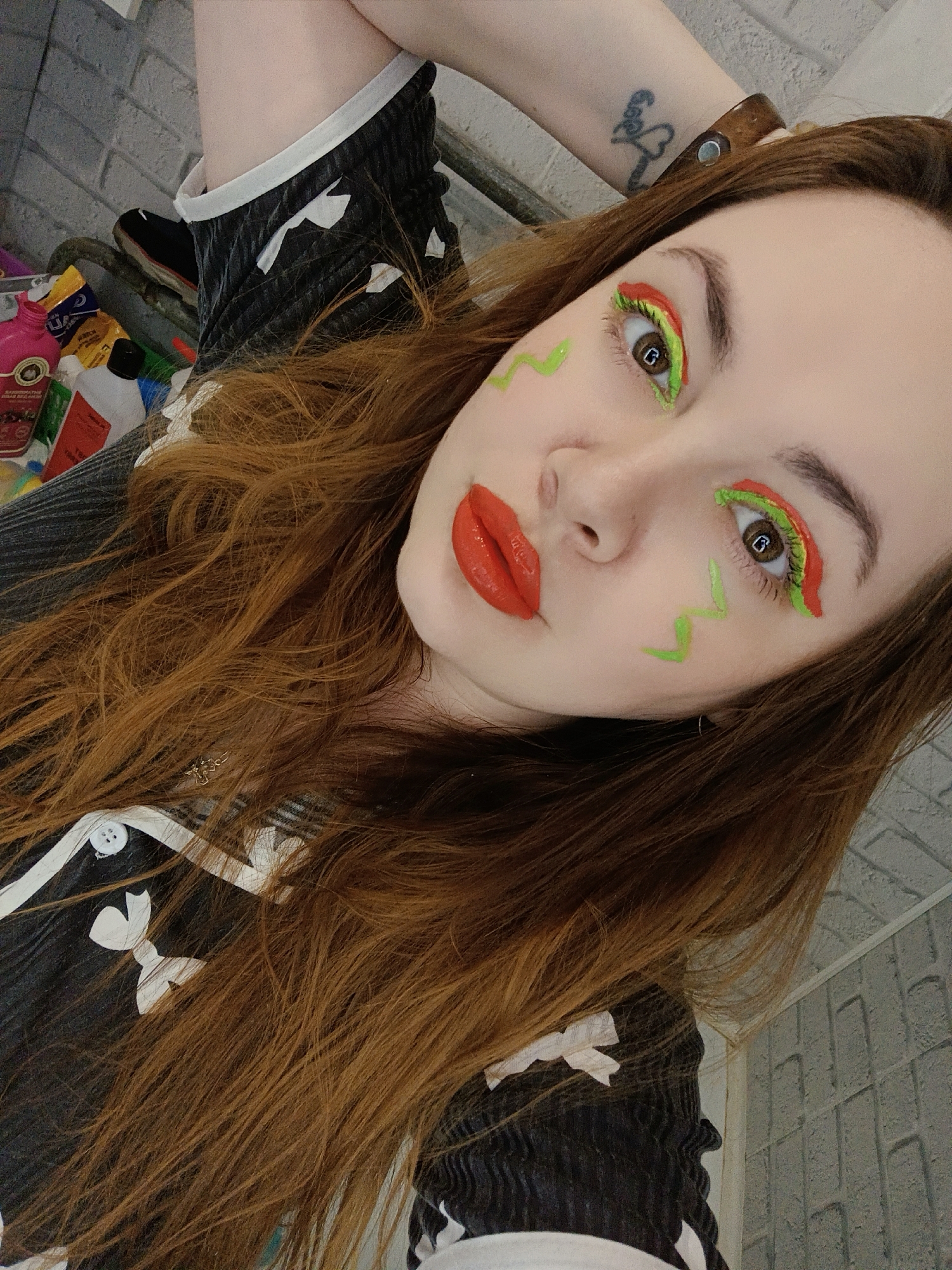 A medium close-up, angled selfie depicts a young woman with long, wavy, reddish-brown hair looking directly at the camera. She features striking, artistic makeup, including vibrant red, glossy lipstick. Her eyes are dramatically adorned with bold green and red eyeshadow or liner, creating a graphic, layered design on her eyelids. Complementing this, green zig-zag lines are drawn on her cheeks. Her eyebrows are dark and well-defined, and her eyes are a warm brown.
She is dressed in a dark garment, likely black, accented with white trim and distinctive white bow-like details on the front. A white button is also visible on her clothing. On her left wrist, she wears a dark, stud-adorned wristband or bracelet, and a small, faded tattoo of a figure holding a heart is visible on her left forearm. A small dark earring is seen in her right ear.
The background suggests an indoor setting that appears somewhat cluttered. To the left, various plastic bottles and containers in shades of pink, yellow, white, and red are visible on what seems to be a shelf or storage unit. A black shoe and a metal bar are also present. The upper right background reveals a grey, textured wall, possibly made of brick or cinder blocks. The lighting is bright, indicative of an indoor photograph. The woman's expression is direct and neutral, emphasizing the creative and playful nature of her makeup. The location context provided for this image is Chaikovsky, Russia.