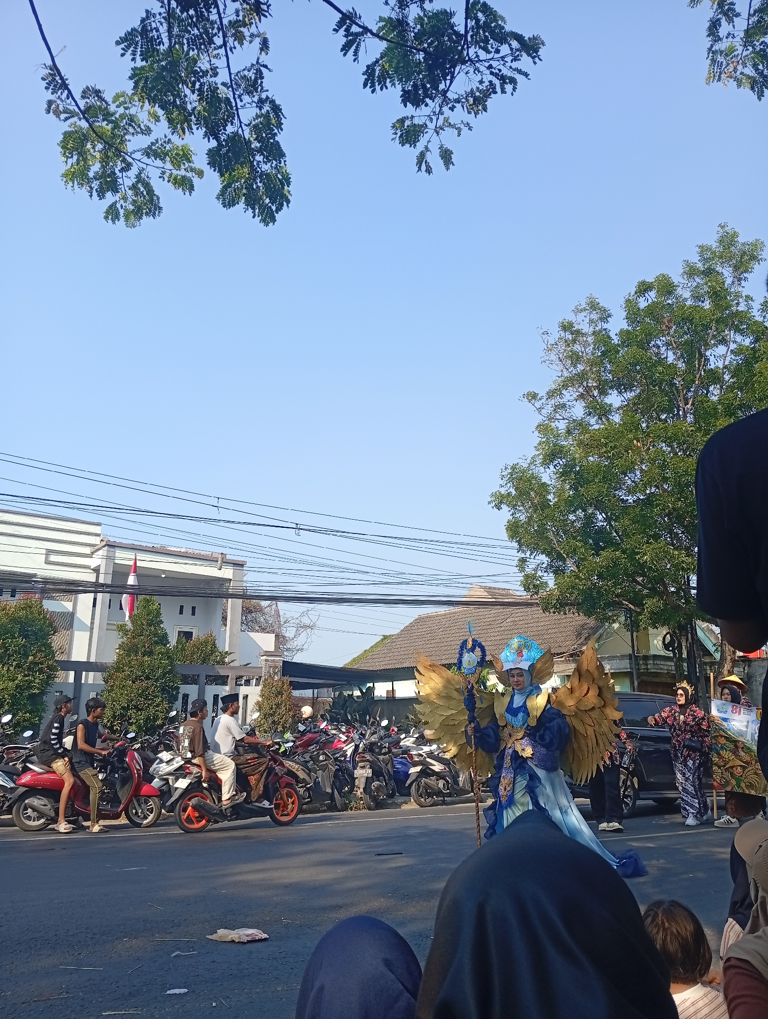 In Bajingjowo, Indonesia, a street scene unfolds under a clear blue sky, with green trees and power lines overhead. The foreground shows the backs of people, some wearing headscarves, watching an event. In the middle ground, a person in an elaborate costume with large golden wings and a blue and gold outfit stands on the street, holding a staff. Behind this performer, a line of motorcycles is parked, and several people are visible. Further back, a building with an Indonesian flag and trees lines the street, with more buildings and trees beyond. The overall atmosphere suggests a parade or cultural event taking place outdoors.