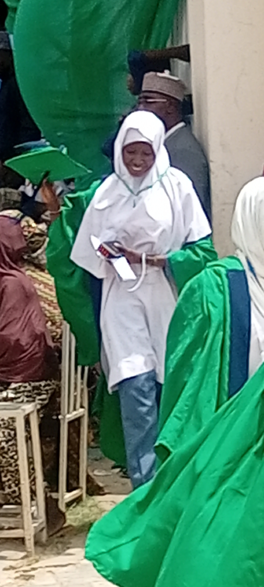 The image shows a woman wearing a white hijab and green graduation gown, holding a red and black item. She is smiling and appears to be at a graduation ceremony. In the background, other people can be seen, including a man wearing a hat and others wearing green gowns. The location is likely Sabon Gari East, Nigeria. There are chairs visible in the background. The ground appears to be paved.