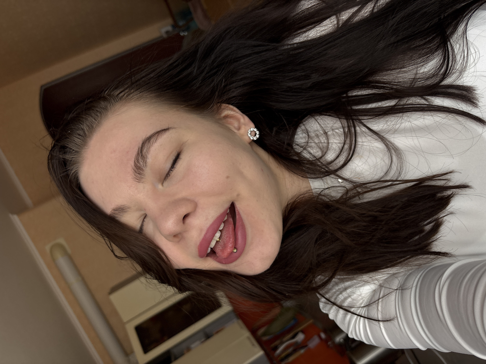 A young woman with dark brown hair is taking a selfie indoors. Her eyes are closed and she is sticking her tongue out, which has a piercing. She is wearing a white top and a round, sparkly earring. The background appears to be a room with furniture. There is no clear evidence to confirm the exact location in the city of Perm, Russia based solely on this image.