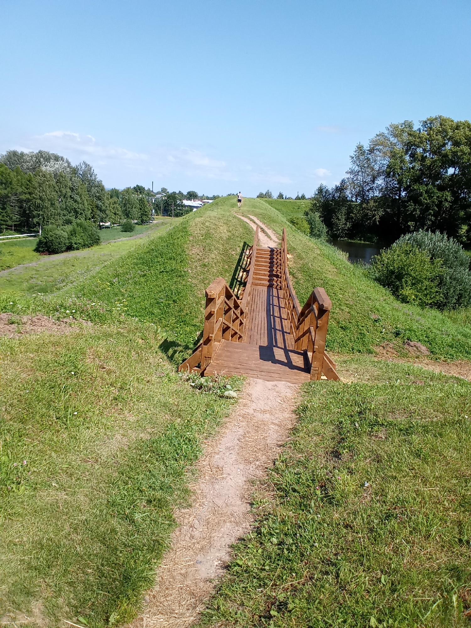 This is a vertical, eye-level shot of a wooden pedestrian bridge crossing a grassy embankment in Belozersk, Russia. The bridge is made of light-colored wood with a planked walkway and railings. It ascends from a dirt path in the foreground and disappears over the crest of the embankment. A single person, visible as a small figure from behind, is walking up the path and onto the bridge. The embankment is covered in green grass with some sparse wildflowers. To the right of the bridge, a small body of water, likely a canal or river, is visible, bordered by dense green trees and bushes. The sky above is clear blue with a few wispy white clouds. The overall scene depicts a sunny day in a park-like or natural setting within the city.