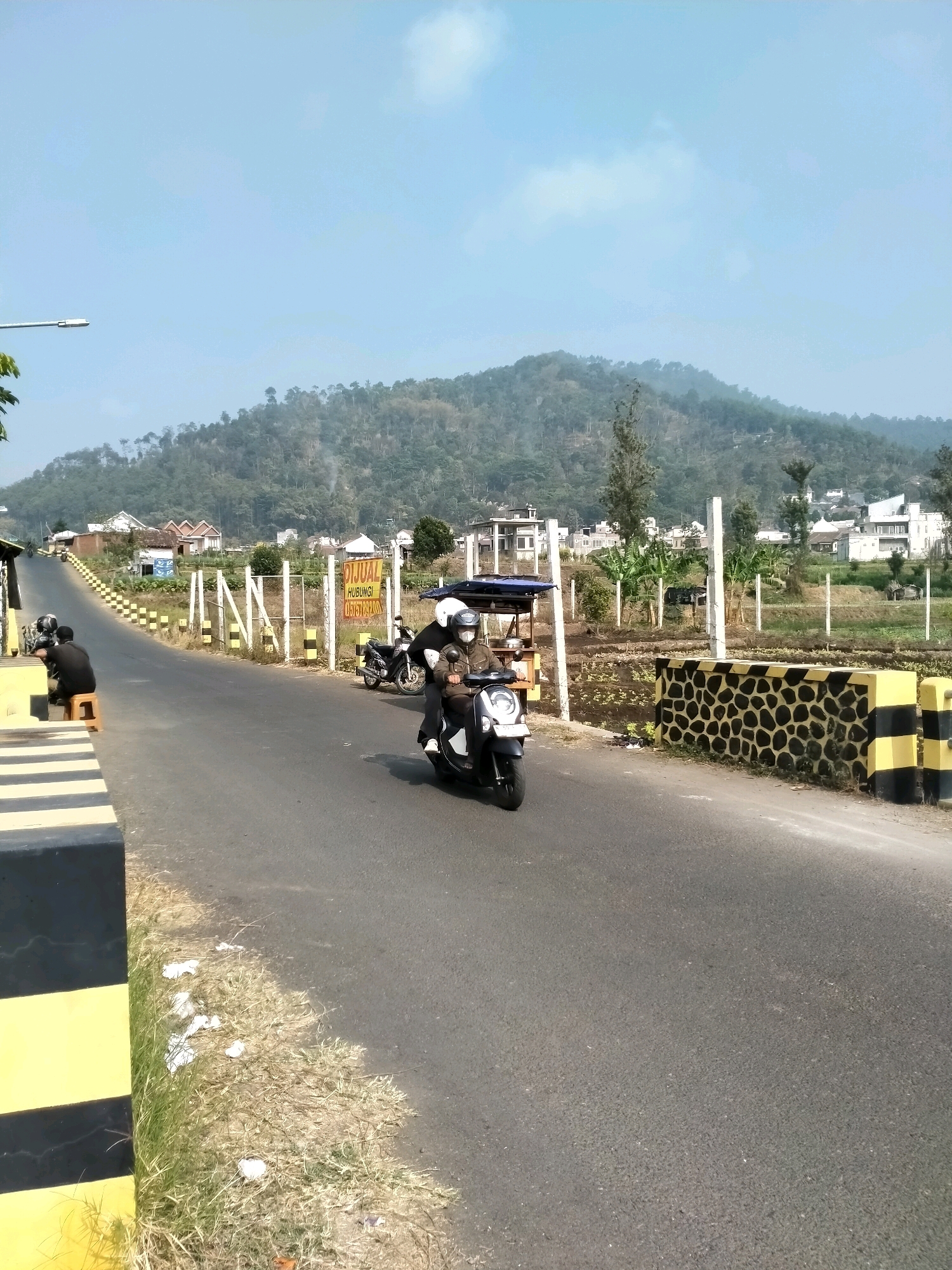 The image depicts a road in a hilly area, likely on the outskirts of Batu, Indonesia. The road curves upwards towards a verdant hillside dotted with houses and trees. A clear sky with sparse clouds hangs overhead. In the foreground, a black and yellow striped barrier lines the side of the road. Two men are seated on stools on the left, near what appears to be a small roadside stall. Further along the road, two people are riding a scooter, with one driving and the other as a passenger. A white motorcycle is parked on the right side of the road, near a fence. A yellow sign with red text, reading "DIJUAL HUBUNGI", is also visible on the right. The environment suggests a semi-rural or suburban setting with agricultural plots visible in the middle ground.