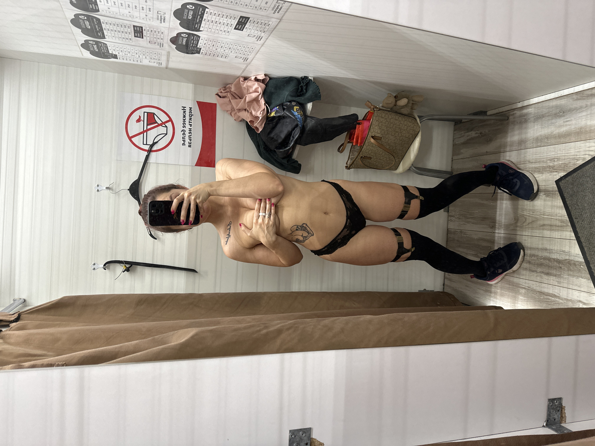 A woman is taking a mirror selfie in what appears to be a dressing room or small retail space. She is wearing black underwear, black stockings with garters, and athletic shoes. She is holding a smartphone and covering her breasts with her hands. The dressing room has a curtain, a clothes rack, and a chair with clothing items and a handbag on it. A sign is visible on the wall with a graphic indicating that wearing underwear is not allowed. The writing on the wall appears to be Russian. Given the writing on the wall, there is no reason to believe the location is Vladimir, Russia.