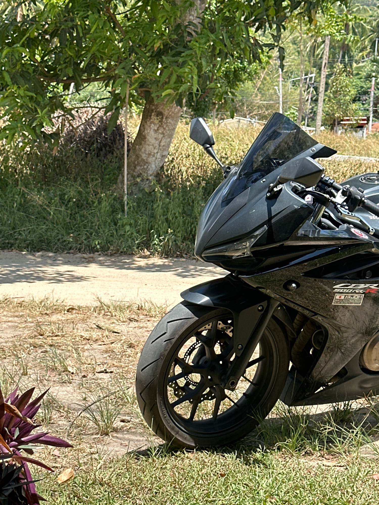 A black Honda CBR 500R motorcycle is parked on a dirt road in a grassy area. The motorcycle is the primary subject, with its front wheel, fairing, and windshield clearly visible. The motorcycle is angled towards the right of the frame, and its black paint is slightly dusty. In the background, there is a tree with green leaves, tall grass, and some palm trees. The ground is a mix of dirt and dry grass. The scene appears to be outdoors, possibly in a rural or semi-rural area. The lighting suggests it is daytime, with sunlight casting shadows. The image was taken in the Ko Pha-ngan District, Thailand.