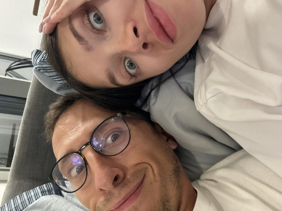 The image captures two individuals, an adult male and an adult female, in a close-up, indoor setting, presumably in Herzliya, Israel. Both appear to be lying down, positioned side-by-side, and facing the camera for a selfie.
The male, situated on the left, has medium-brown hair, a lightly stubbled beard, and visible wrinkles on his forehead. He wears black-framed glasses with reflections, through which his left eye is visible, appearing enlarged and looking upwards. His mouth is closed in a slight, asymmetrical smirk, with the left corner downturned. He is wearing a plain white t-shirt.
The female, on the right, has long, dark hair. Her eyes are wide open, directed straight at the camera, conveying a surprised or playful expression. She has prominent eyelashes and full, light pink lips that are slightly pursed. Her right hand, showing manicured nails with a light polish, is partially visible on her forehead/hairline. She also wears a plain white t-shirt.
Both subjects are resting on light gray bedding, with a light blue and white striped fabric visible behind the male's head. The background is indistinct and out of focus, indicating an indoor domestic environment.
