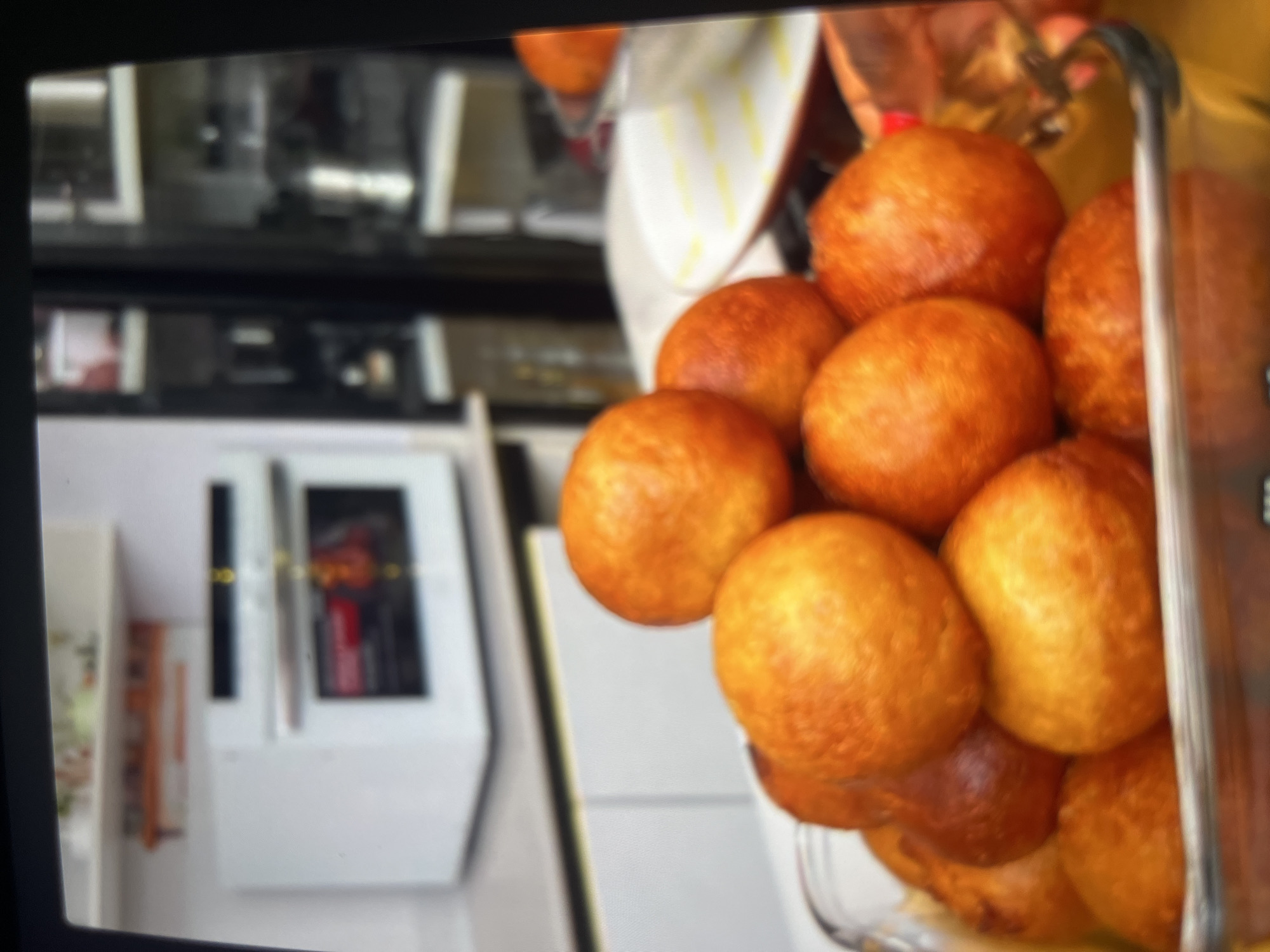 The image shows a close-up of a pile of golden-brown, spherical fried dough balls, likely puff-puffs, in a clear glass dish. The dough balls are clustered together, with some overlapping. In the background, a white oven is visible on a countertop, and to the right, a refrigerator with glass doors displays shelves stocked with various items. The overall setting appears to be a kitchen. The image is grainy and appears to have been taken from a screen, possibly a mobile phone or television. The location is specified as the city of Abuja, Nigeria.