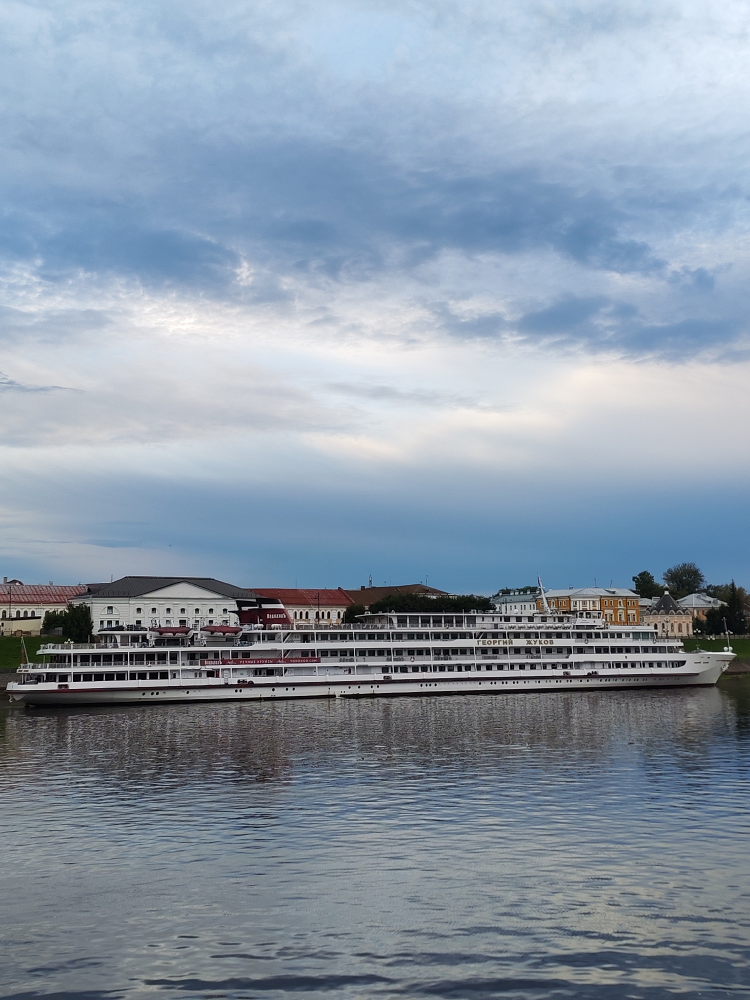A large, white, multi-decked passenger ship, named "ГЕОРГИЙ ЖУКОВ" (Georgy Zhukov) in Russian Cyrillic lettering, is docked on a wide, calm river. The ship is positioned horizontally across the frame, with its reflection visible on the water's surface. Behind the ship, on the riverbank, are several buildings, including a long white building with a dark roof, and a building with a yellow facade and red roof. The sky above is filled with dramatic, cloudy formations, with patches of lighter sky suggesting the presence of the sun. The scene appears to be an urban waterfront in Bol'shie Mkhi, Russia, likely in the late afternoon or early evening, given the lighting and cloud conditions. No people are discernible in the image.