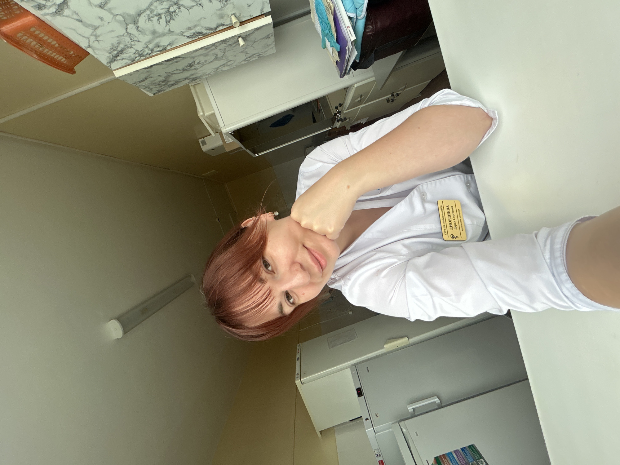 A young woman with reddish-brown hair and fair skin is posing for a selfie. She is wearing a white medical coat with a name tag, which appears to be written in Russian. The name tag reads "ДИОРДИЕВА Дарья Сергеевна." Her right arm is bent and her fist is touching her chin. She is looking directly at the camera with a slight smile. The background appears to be a medical or office setting. A refrigerator and some office furniture are visible. It is likely in Фоки, Russia.