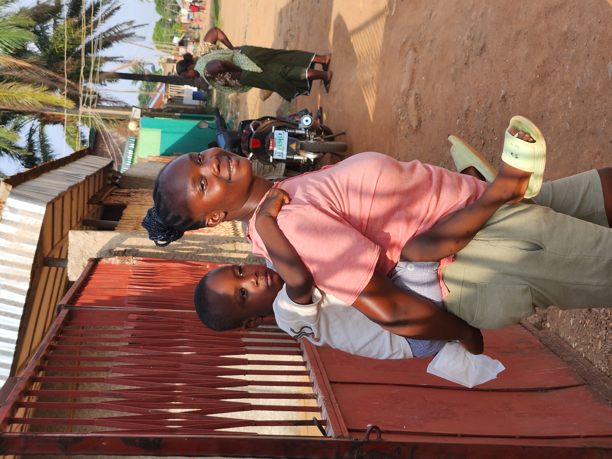 In a street scene in Sabon Tasha, Nigeria, a woman is holding a young child on her back. The woman is wearing a pink t-shirt and green pants, and the child is wearing a white t-shirt. They are both looking at the camera with smiles. In the background, there are palm trees, buildings, and a motorcycle. The ground is dusty, and the sun is shining, suggesting it is daytime. The overall mood of the image is warm and joyful.