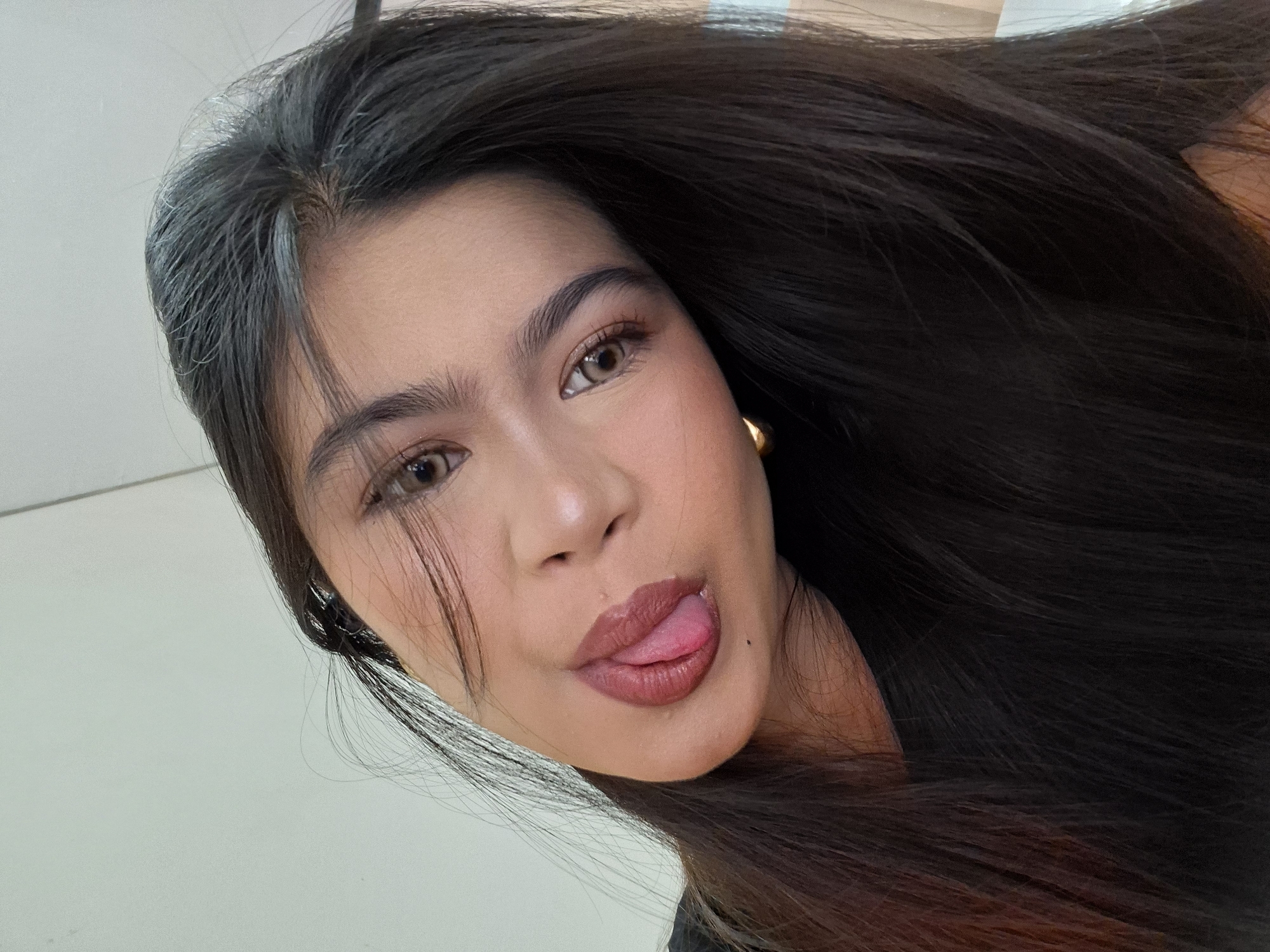 A close-up, eye-level selfie of a young woman with long, dark hair, tilted head and her tongue sticking out. She is wearing dark clothing and a gold earring. The background is a plain, light-colored wall, suggesting an indoor setting, possibly a room in Kuta, Indonesia, given the user's prompt. The lighting is bright, casting a soft glow on her face.