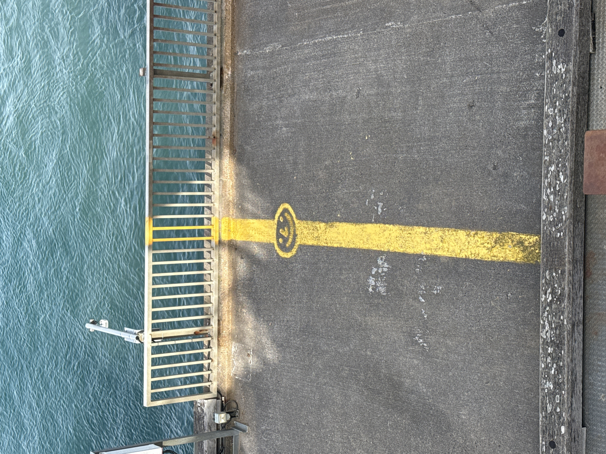 Captured from an elevated perspective, the image presents a section of a pier or walkway, adjoining a body of water. A metal railing lines the left edge of the walkway, with turquoise water visible beyond it. A yellow stripe runs horizontally across the grey asphalt surface, featuring a circular design with the number '3' inscribed within it. To the right, a weathered wooden beam marks the edge of the walkway. The location is Unknown, Unknown.