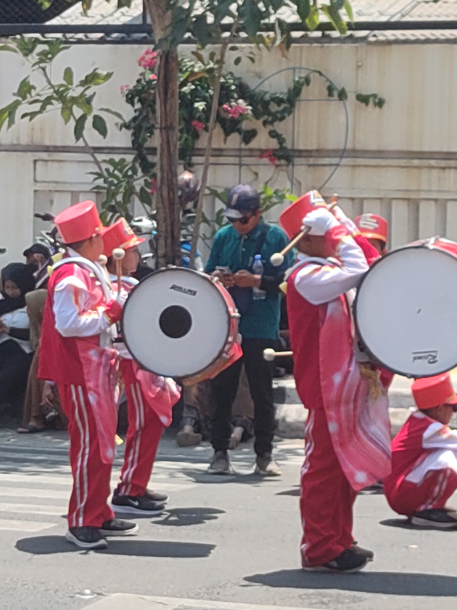 In a sunlit outdoor scene on a paved street in Ngasem, Indonesia, a group of individuals in marching band uniforms are visible. The primary subjects are two young people wearing bright red uniforms with white accents, including tall red hats, and white gloves. They are holding large bass drums, one on each side of the frame, and appear to be playing or preparing to play them with drumsticks. The drum on the left is white with a black center and has the word "ROLLINS" visible on it. The drum on the right is also white.
Behind the drummers, other individuals are present. A person wearing a teal shirt and sunglasses is standing and looking at their phone. Further back, more people are seated or standing, some wearing headscarves. The background consists of a pale wall, a fence, and greenery from trees with pink flowers. The ground is a crosswalk with visible white stripes. The overall atmosphere suggests a daytime parade or performance.