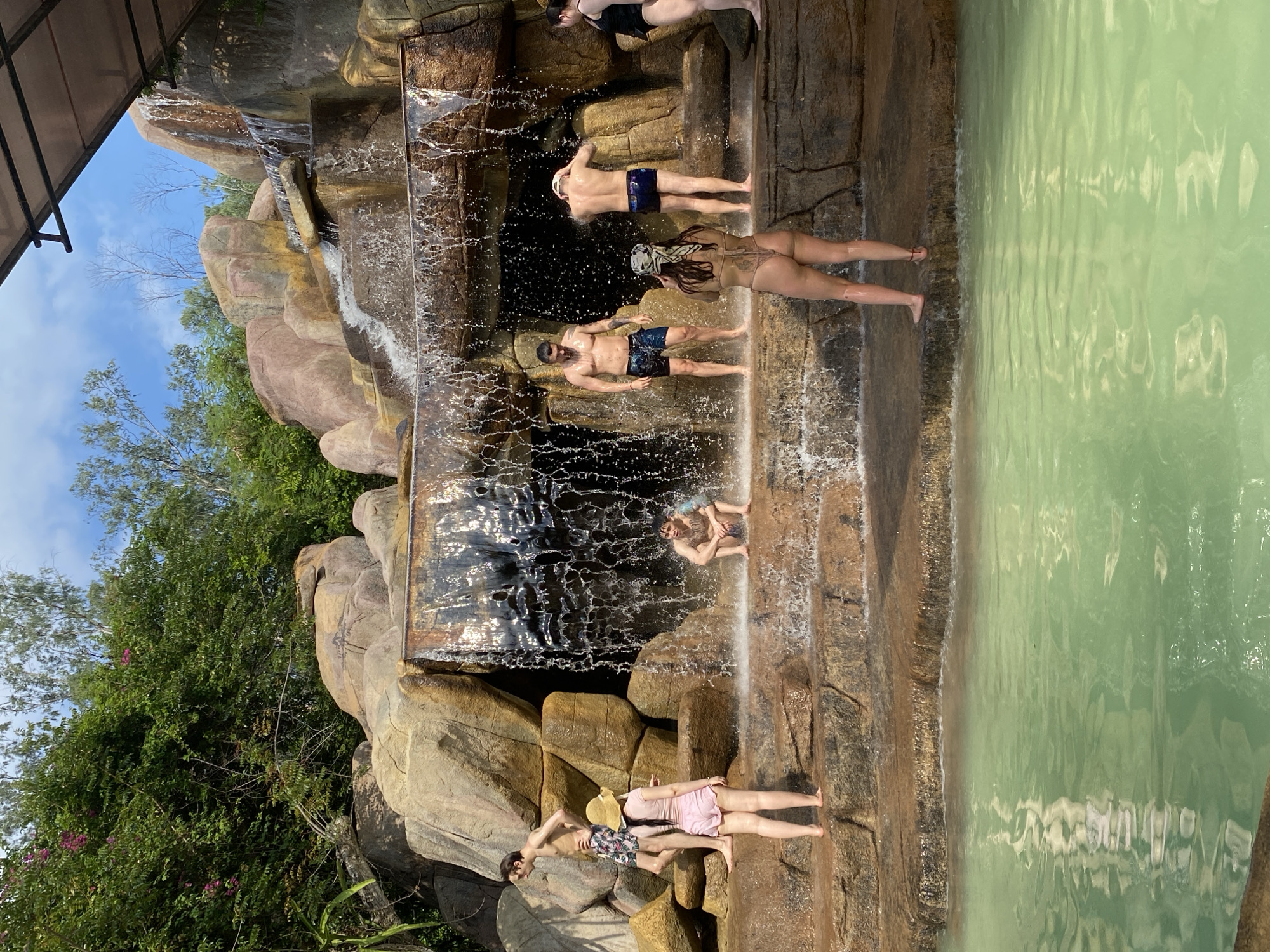 In Tay Nha Trang Ward, Vietnam, a group of people are enjoying a man-made waterfall in a recreational area. The waterfall cascades over several tiers of textured, faux-rock formations, creating a misty atmosphere. The water plunges into a clear, jade-green pool in the foreground. Several individuals are interacting with the waterfall, some standing beneath the falling water, others on the rocky ledges. One person sits on a lower ledge, partially submerged in the water. The surrounding environment appears lush with greenery and trees, and the sky above is partly cloudy. The scene depicts a relaxing day at a water attraction.