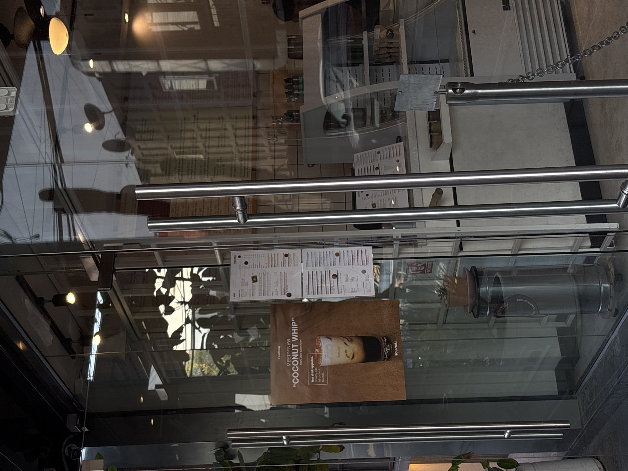 The image depicts a storefront or entrance, likely in a restaurant or café, with glass doors and metal handles. Several signs are visible on the glass: menu items, including one prominently advertising a "Coconut Whip" beverage from SOULVALI. Reflections on the glass show a building exterior and vegetation, suggesting an outdoor setting. The overall environment appears urban and commercial, possibly in Puebla, Mexico.