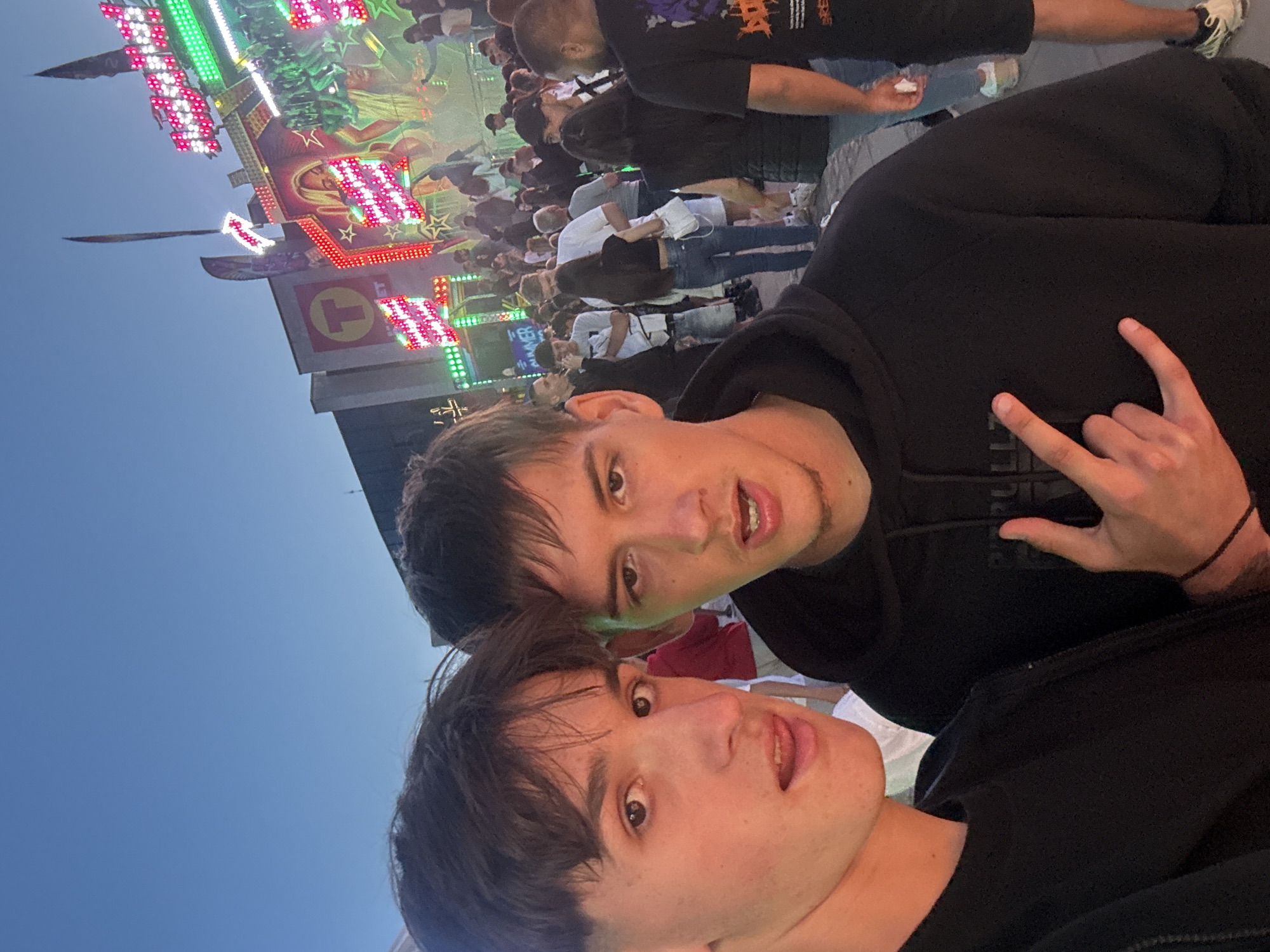 Two young men pose for a selfie at a fairground. The man in the foreground wears a black shirt, while the other, slightly behind, dons a black hooded sweatshirt and flashes a "rock on" hand gesture. Behind them is a crowd of people and a lit-up carnival ride, and a blue sky is visible in the background. The location may be Bozhurishte, Bulgaria.