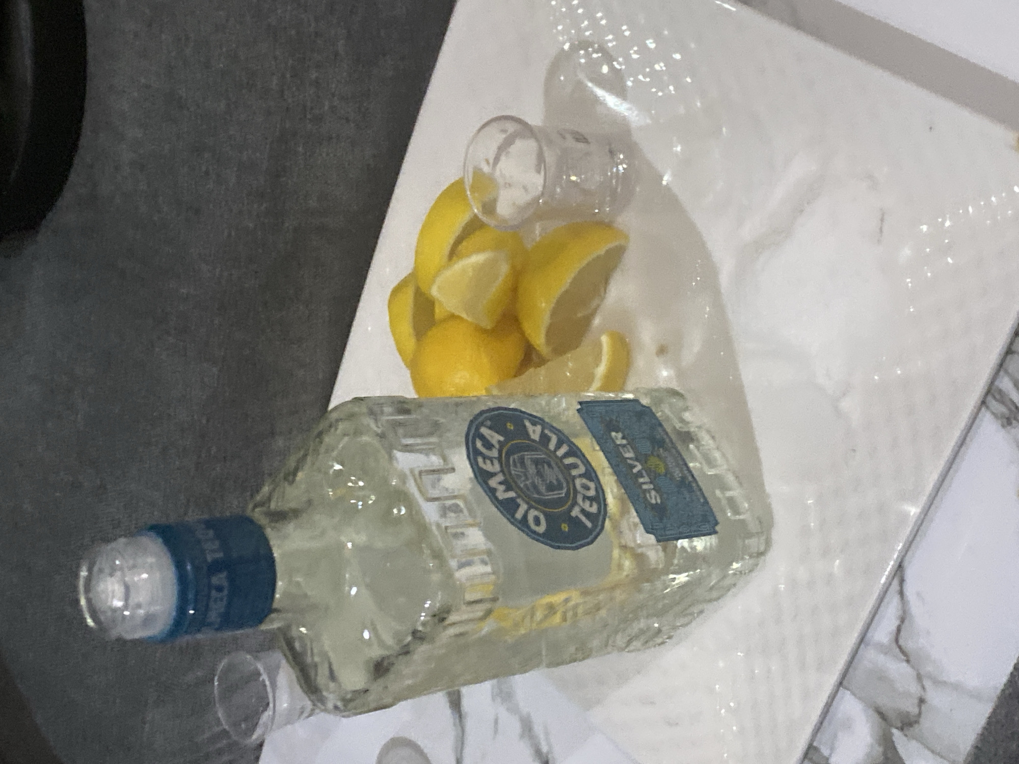 The image captures a still life arrangement centered around a bottle of tequila, accompanied by lemon wedges and shot glasses, set on a white tray. The scene is indoors, located in Iddo Tudunwada, Nigeria, though no specific visual cues within the image indicate this geographical context.
The main subject is a clear glass bottle of "OLMECA TEQUILA SILVER," lying horizontally on the white rectangular tray. Its label, primarily blue and gold, clearly displays the brand name "OLMECA TEQUILA" and the product type "SILVER." Further text, "MADE WITH BLUE AGAVE," is also discernible on the label.
Clustered near the bottle on the tray are numerous bright yellow lemon wedges, indicating preparation for tequila shots. Two small, clear shot glasses are present: one lies on its side amidst the lemon wedges on the tray, and another is partially visible at the bottom left corner of the frame, also on its side.
The white tray has a slightly reflective surface. It rests on a dark gray, subtly textured surface, likely a table or countertop. To the right of the tray, a distinct white and gray marbled surface is visible, possibly a floor or another countertop. The lighting suggests an indoor setting, with reflections visible on the glass bottle, shot glasses, and the tray. No people are depicted, and no specific activity, time of day, or emotions are explicitly shown, but the arrangement strongly implies a social drinking event involving tequila.