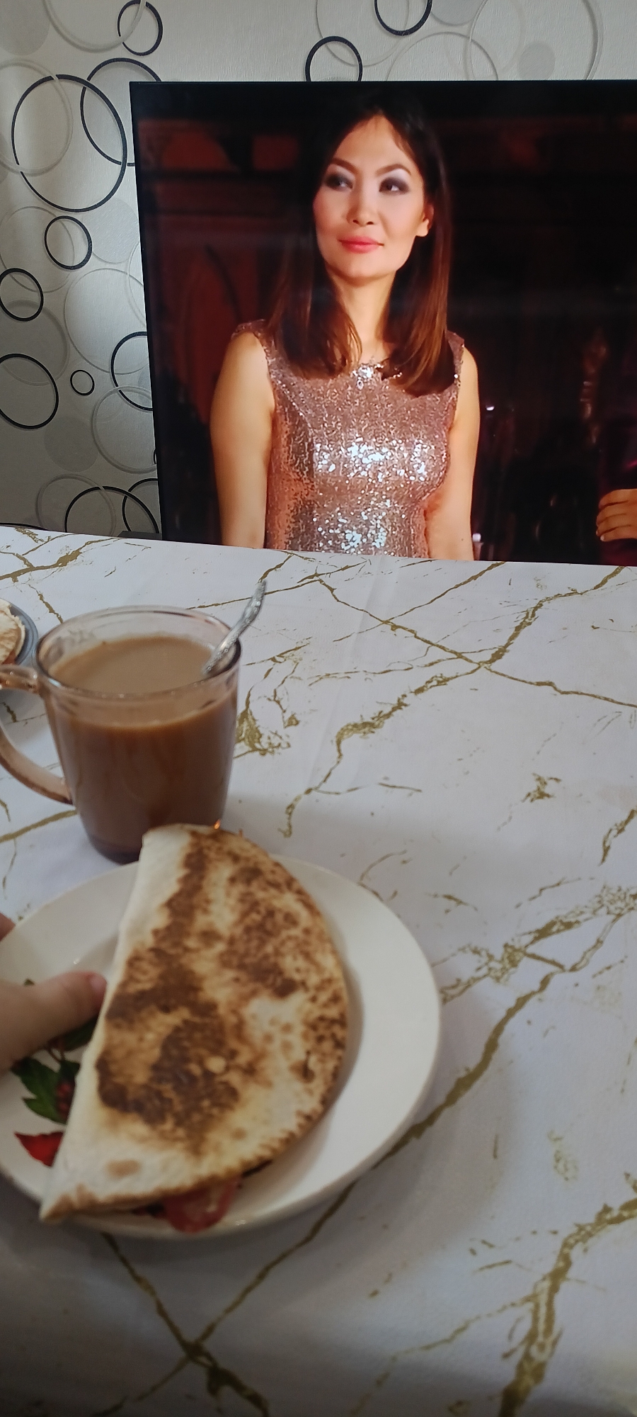 A close-up shot shows a table with a meal and a television in the background. The television screen displays a woman wearing a sparkling pink dress. On the table, a mug filled with coffee and a piece of quesadilla are visible. The table is covered with a white tablecloth featuring a gold marbled pattern. The background shows a wall with a circular pattern.