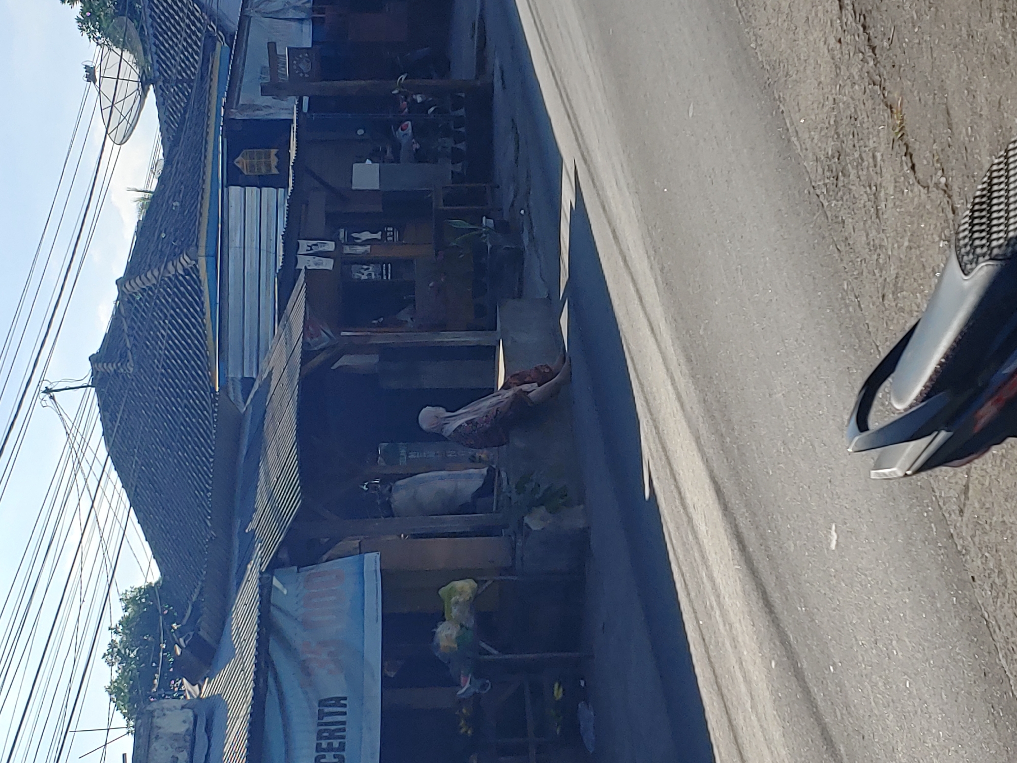 In Kembang Kerang, Indonesia, a lone woman wearing a headscarf and a patterned dress sits on a low concrete ledge in front of a small shop. The shop has a corrugated metal roof and a sign with the word "CERITA" visible. To the left of the shop, a table displays produce, including bananas. Behind the woman, a large white bag is leaning against the wall. The scene is set along a paved road, with a motorcycle's rear seat visible in the foreground. The sky above is clear and blue, with a network of overhead wires crisscrossing the frame. A satellite dish is mounted on a rooftop in the background.