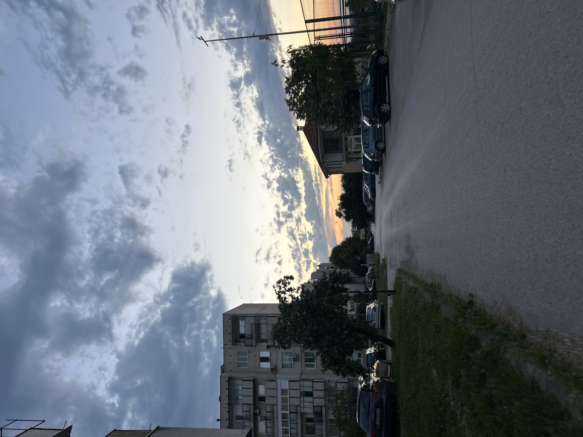 The image depicts an urban street scene at dusk. The sky is filled with dramatic clouds, with hints of orange and yellow from the setting sun visible near the horizon. On the left side of the street, there is a multi-story apartment building with balconies and windows. A tree stands in front of the building, casting a shadow on the grassy area beside the sidewalk. Several cars are parked along the street.
On the right side of the street, there is a building with a red-tiled roof and a sports field with a fence behind it. More trees line this side of the road. The street itself is paved and appears to be empty of pedestrians in the foreground. The overall impression is a quiet evening in a residential area.