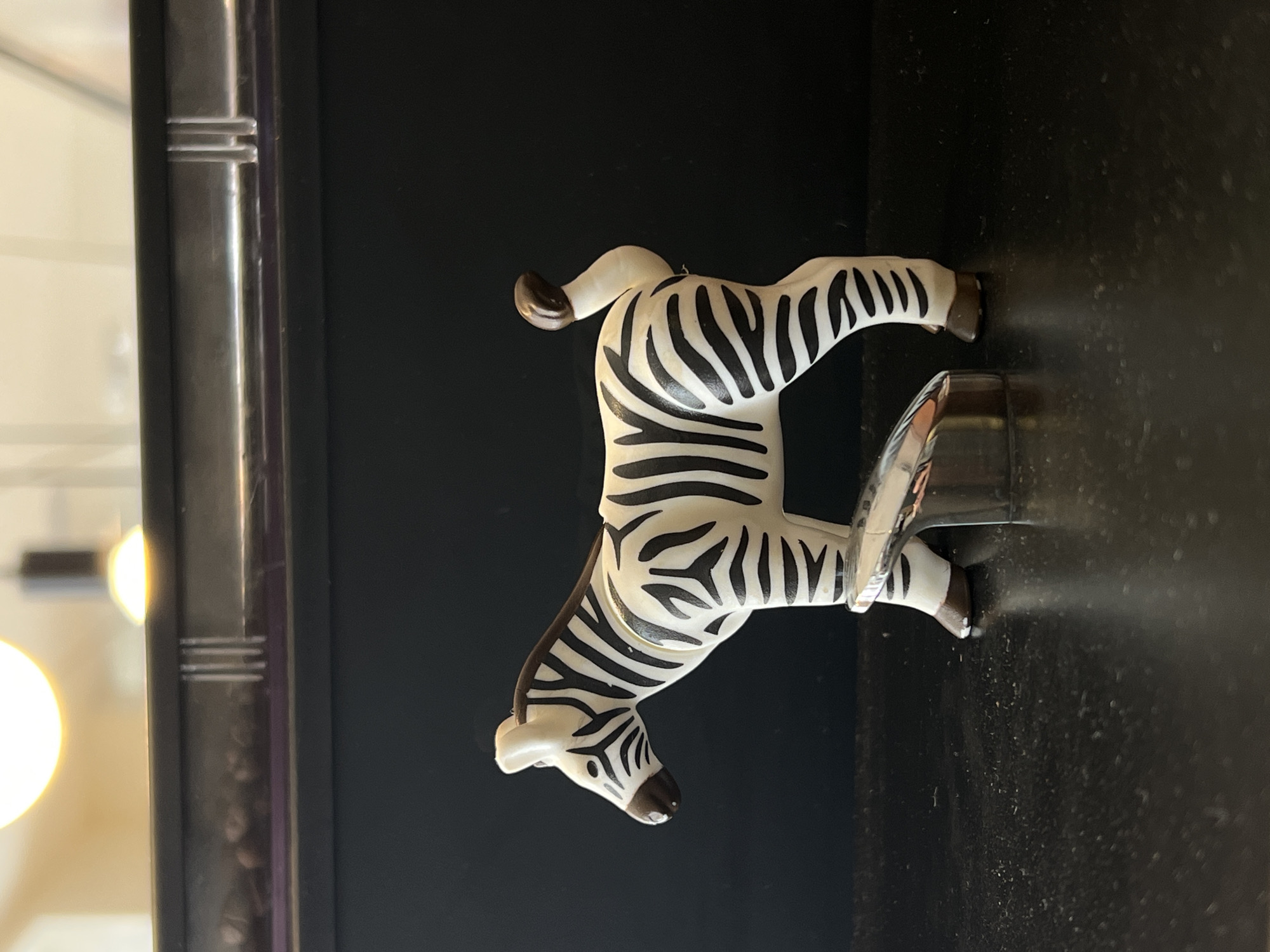 A small, plastic zebra figurine, predominantly white with black stripes, stands on a dark, reflective surface. The figurine is positioned in profile, facing left. Its head is down slightly, as if grazing. A metallic, cylindrical object, possibly a lighter or a cap, is placed in front of its front legs, partially obscuring them. The background is dark, with a horizontal element above, featuring a blurred bright light source and some indistinct objects. The overall scene appears to be indoors, on a shelf or tabletop. There is no discernible connection to Tel Aviv, Israel.