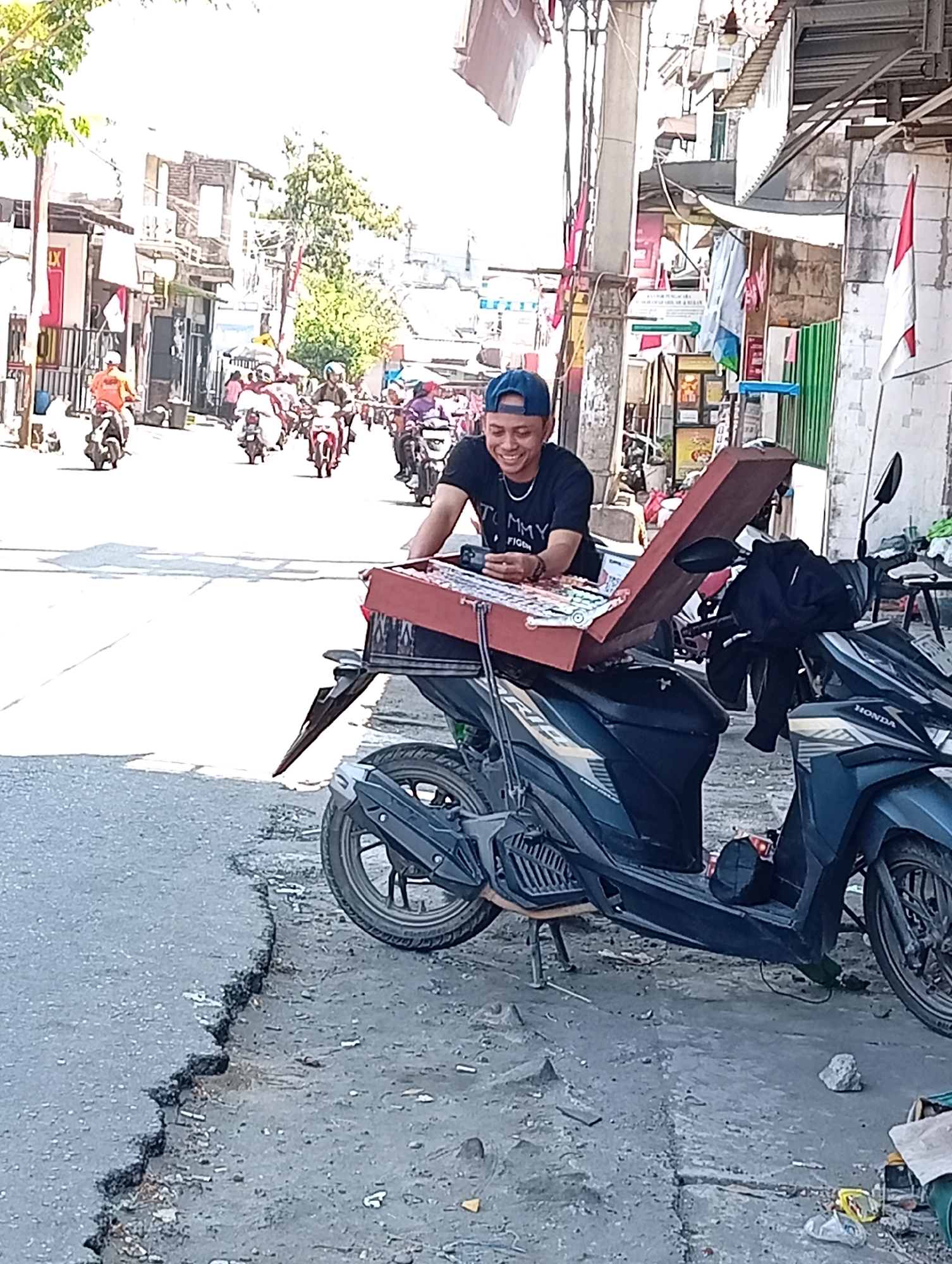 A man wearing a blue baseball cap and a black t-shirt is smiling while sitting on a dark-colored motorcycle. He is looking down at a red box containing various items, possibly merchandise, placed on the motorcycle's seat. The man's left hand rests on the box. The motorcycle is parked on a cracked asphalt road. In the background, a busy street is visible with several other motorcycles and pedestrians. Buildings line both sides of the street. The overall setting suggests a street market or a bustling urban area in Surabaya, Indonesia, on a bright, sunny day.