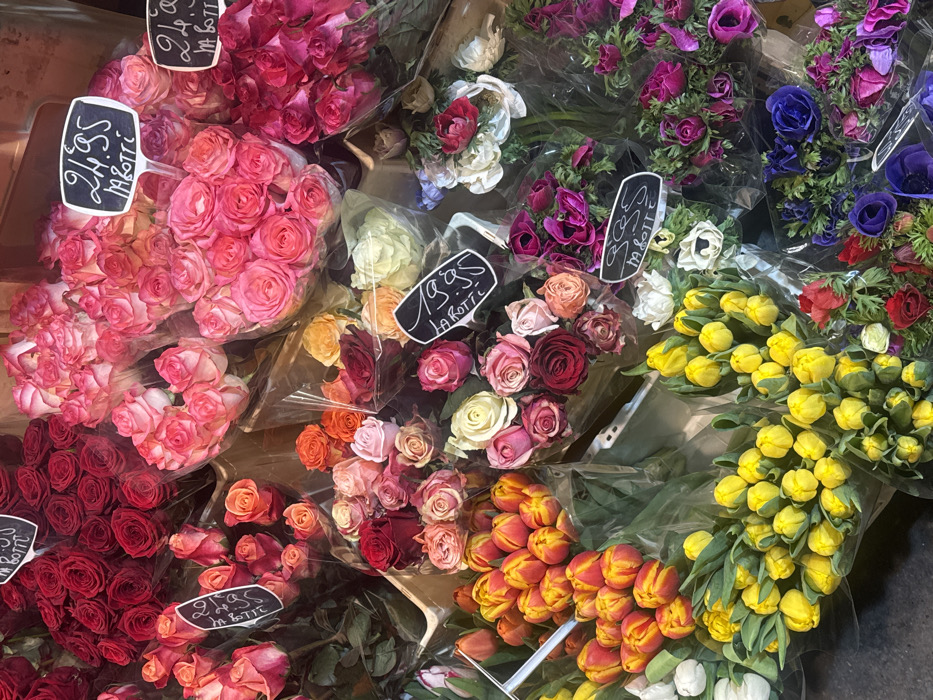 The image displays a dense, top-down view of numerous flower bouquets arranged for sale in what appears to be a market stall or flower shop. The primary subjects are fresh cut flowers, predominantly roses and tulips, bundled and wrapped in clear plastic sleeves.
Roses are present in various colors, including deep red, vibrant pink, two-tone pink and white, orange, peach, and maroon. Several bouquets of yellow tulips with prominent green stems are visible in the lower section, alongside bundles of red and orange tulips. Other floral arrangements include purple, blue, and white flowers resembling anemones or ranunculus.
Interspersed among the bouquets are small, black, chalkboard-style signs with white handwritten text. Visible pricing includes "21.95 M.BOTTE" on two signs in the upper right quadrant, "19.95 LA ROTTE" on a sign in the mid-left, and "9.95 M.BOTTE" on a sign in the mid-right. A partial sign showing "19.95 LA ROTTE" is in the lower right. The flowers are arranged vertically, suggesting they are held in unseen containers such as buckets. The background is dark and indistinct, likely the floor or display surface of the stall. No individuals are present in the frame.
The location is Unknown, Unknown.