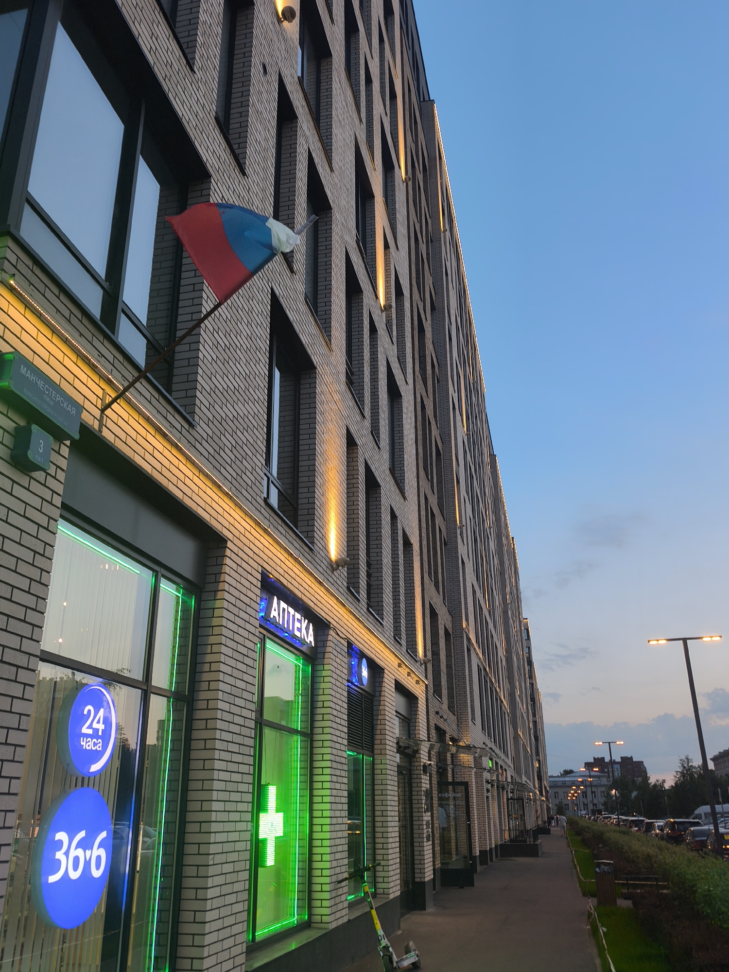 A long, modern building with a light brick facade stretches diagonally across the frame, receding into the distance. The building has numerous rectangular windows, some reflecting the blue evening sky. Warm yellow lights are embedded along the facade, creating a line of illumination. On the left side of the building, a sign reads "МАНЧЕСТЕРСКАЯ" and "3", indicating Manchester Street, house number 3. Below this, a store window is brightly lit with green neon accents, displaying circular signs reading "24 часа" (24 hours) and "36.6". Another store sign, "АПТЕКА" (Pharmacy), is visible with neon lighting. A Russian flag is mounted on the building near the top left. The sidewalk in front of the building is paved, and a green hedge runs alongside it. A streetlamp with multiple lights is visible on the right side of the frame, and parked cars line the street in the distance, suggesting an urban environment at dusk. A green and white scooter is parked on the sidewalk in the foreground.