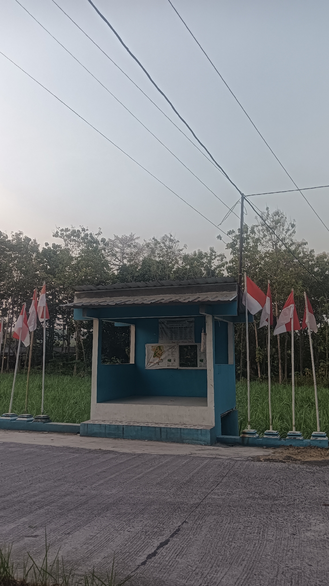 In the city of Srawung, Indonesia, a small, blue-painted guard post stands prominently. The post has a grey roof and a light-colored base. In front of the post, several Indonesian flags, red and white, are planted on poles. Behind the guard post, a dense line of trees forms a natural backdrop. The ground in the foreground is paved with a dark grey asphalt-like material, showing parallel lines from its construction. The sky above is a hazy pale grey, suggesting either dawn or dusk. Power lines are visible diagonally across the upper portion of the image.