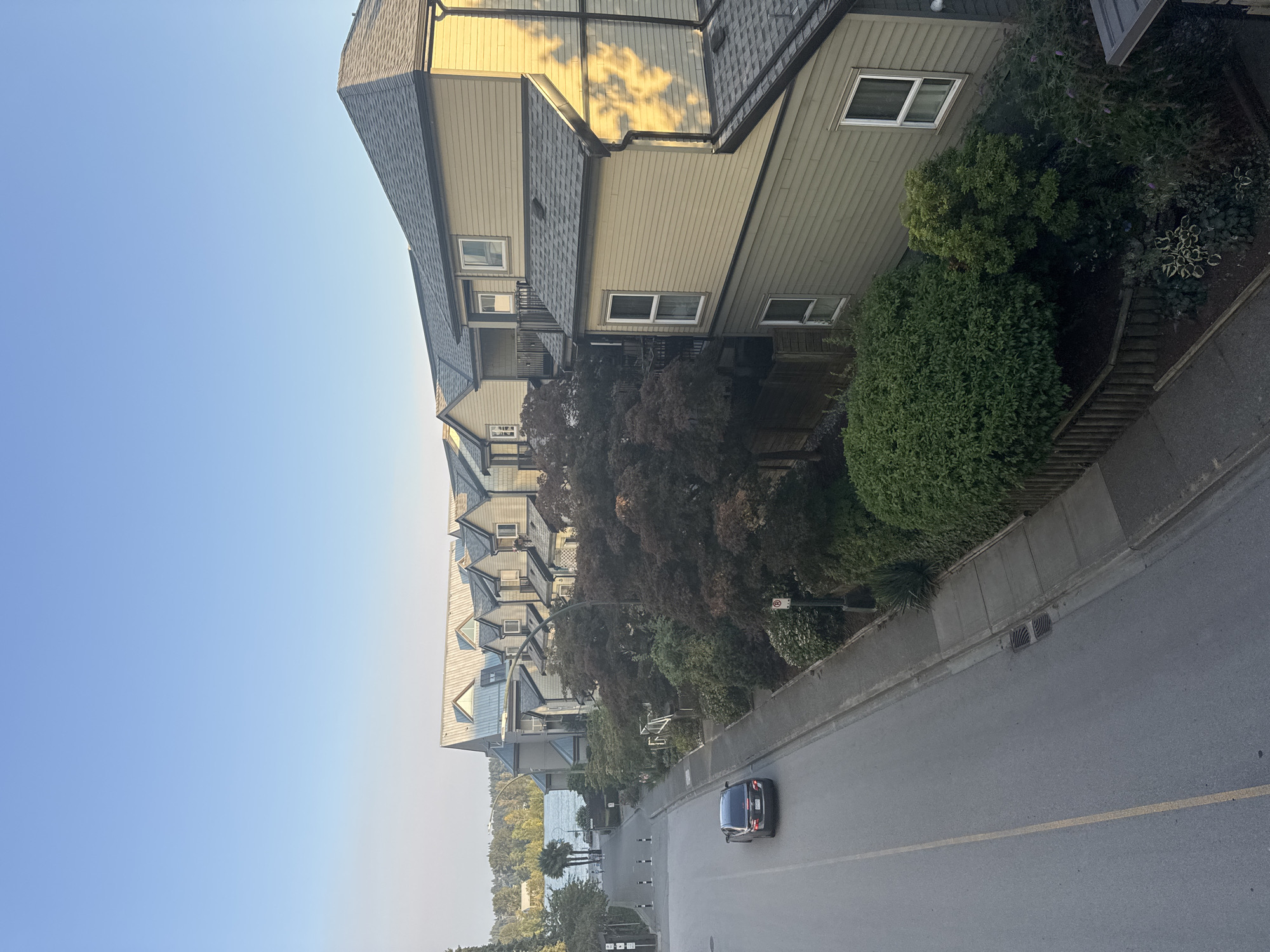 A high-angle shot captures a street scene in New Westminster, Canada, with a row of beige townhouses lining the right side of the frame. A dark sedan is driving away from the camera on the left side of the street. The townhouses have dark grey pitched roofs and multiple windows. Lush green trees and bushes are planted in front of the townhouses. A sidewalk runs along the street, and a few streetlights are visible. In the background, a body of water, possibly the Fraser River, is visible, with trees and buildings on the far bank under a clear blue sky. The sun casts shadows on the side of the townhouses.