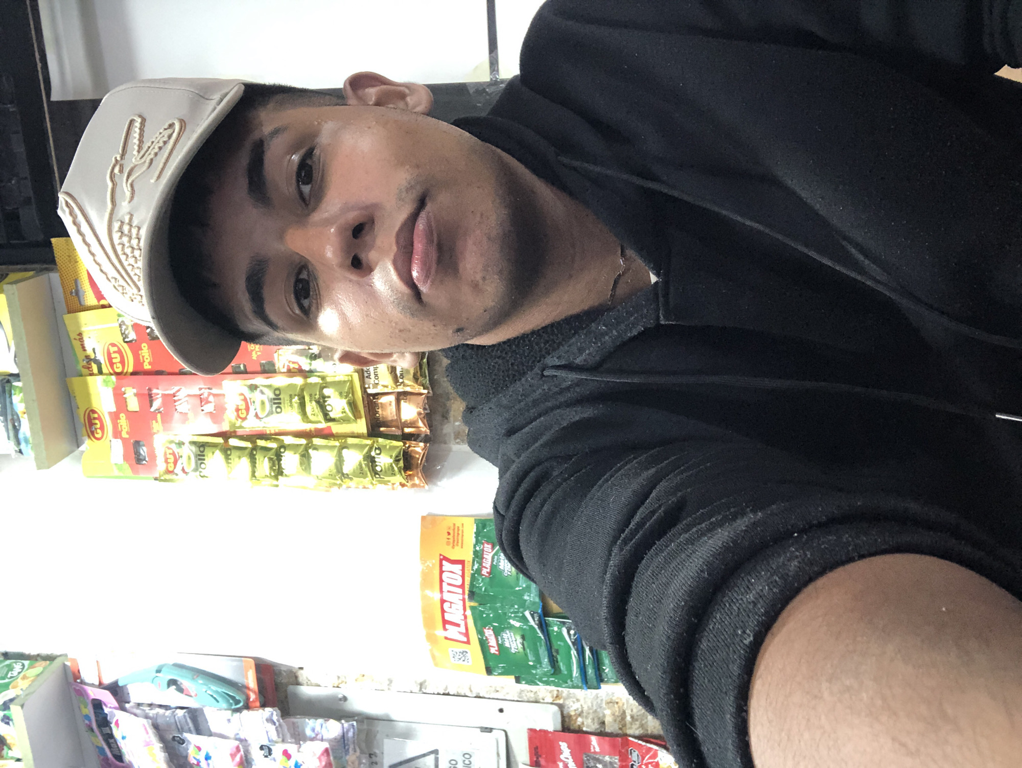 A person with tan skin, dark hair, and wearing a light-colored cap with a crocodile logo is the main subject of the image. They are wearing a black hooded sweatshirt. The background reveals a store shelf stocked with various products including seasoning packets, Plagatox insecticide, and assorted small items. The presence of the insecticide and the labels on the seasoning packets suggest the location might be a small, possibly family-owned business in Venezuela. Although the specific city, Juan Griego, is mentioned, there is no visual confirmation within the image to confirm it is in this location. The image is taken at an angle, suggesting it might be a selfie.