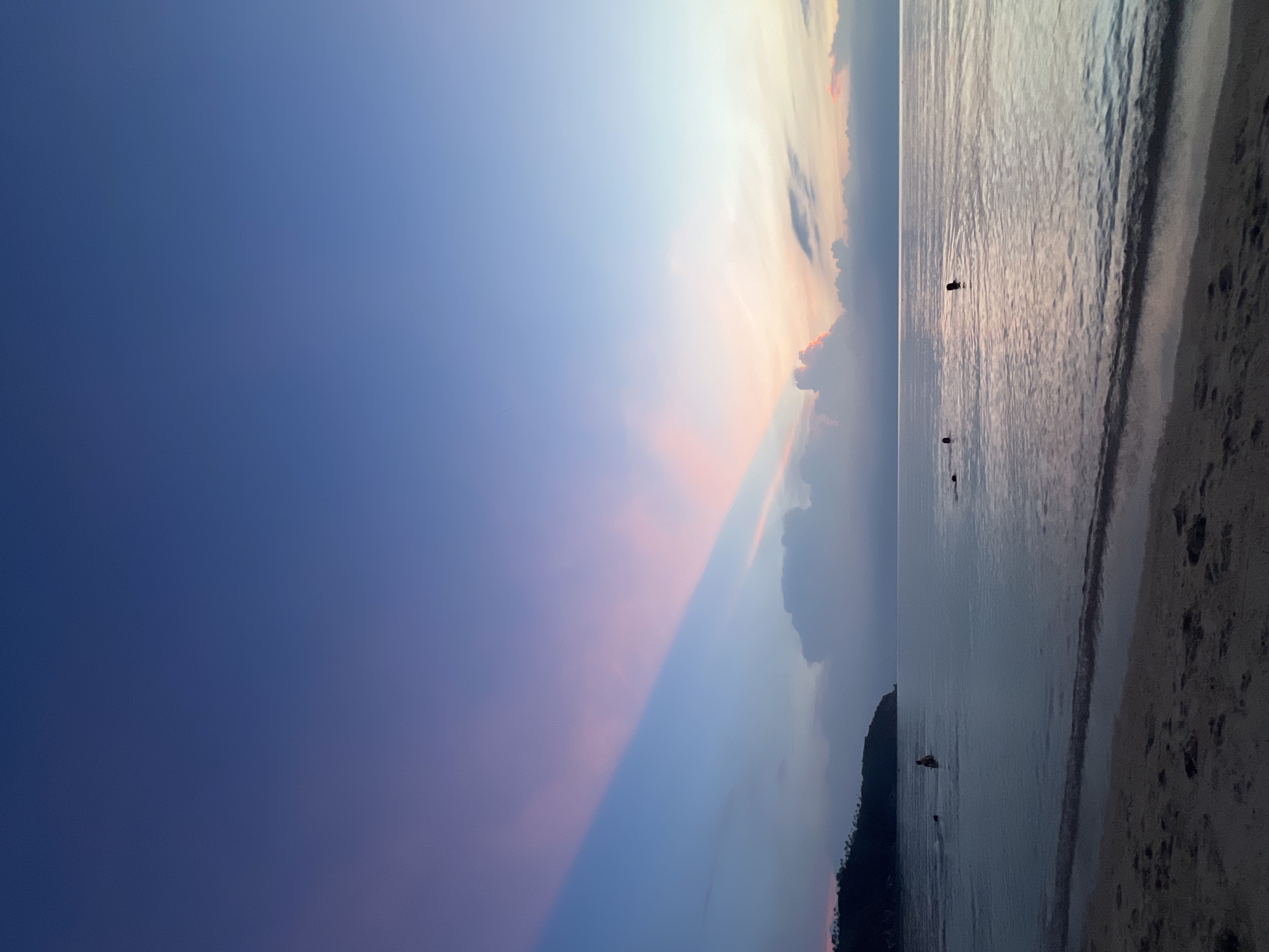 A vertical, low-angle shot captures a tranquil sunset scene on a beach in Ko Pha-ngan District, Thailand. The sky transitions from a deep blue at the top to lighter hues of purple and pink as it nears the horizon, with a dramatic band of pinkish-purple clouds streaking diagonally across the upper left portion of the frame. The calm ocean reflects the sky's colors, with gentle waves lapping at the sandy shore in the foreground. Several people are visible in the water, appearing to swim or wade, their forms silhouetted against the soft light. In the distance, a tree-covered headland juts out into the sea on the left side of the image. The overall atmosphere is serene and peaceful.