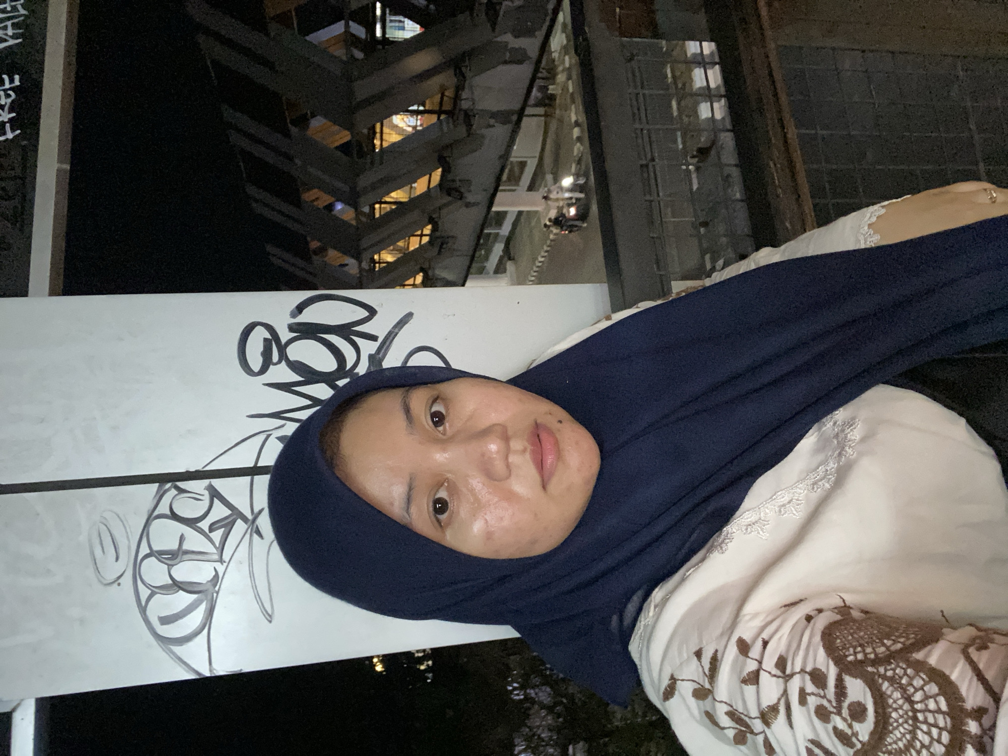 A selfie of a woman wearing a dark blue hijab and a cream-colored top with brown embroidered details. She is positioned slightly to the left of the frame, looking directly at the camera. The background is a night scene with an elevated structure featuring angular supports, illuminated from within. Graffiti is visible on a white pillar to the left of the woman. In the distance, a street is visible with a few people and motorcycles. The setting appears to be an urban environment in Banjarbaru, Indonesia.