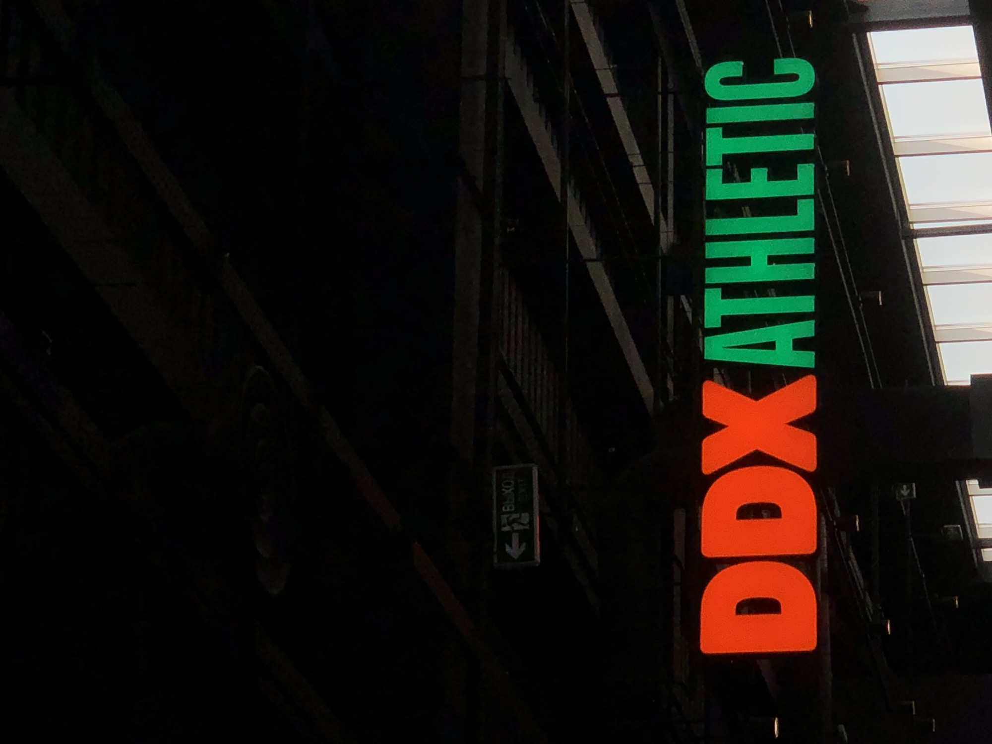The media file displays a nighttime or dimly lit indoor scene featuring a prominent, vertically oriented illuminated sign for "DDX ATHLETIC." The top portion of the sign, "ATHLETIC," is rendered in bright green capital letters, while the bottom portion, "DDX," is in vibrant orange capital letters. The sign glows brightly against a predominantly dark background, which reveals hints of industrial-style architectural structures, likely girders or beams, behind and around it. These structures appear dark and form abstract patterns due to the low light.
To the left of the main sign, further in the dark background, a smaller emergency exit sign is visible. This sign is also illuminated and features white text against a dark background, reading "ВЫХОД" (Cyrillic for EXIT) above "EXIT" in English, with a downward-pointing arrow indicating the direction.
The overall setting appears to be a modern building, possibly a shopping complex or a fitness center itself, located in Krasnoyarsk, Russia, as indicated by the dual-language exit sign and the contemporary branding. No people or activities are visible, emphasizing the static nature of the architectural elements and signage. The bright illumination of the signs strongly contrasts with the dark surroundings, suggesting the image was captured during the evening or within a structure with minimal ambient lighting.