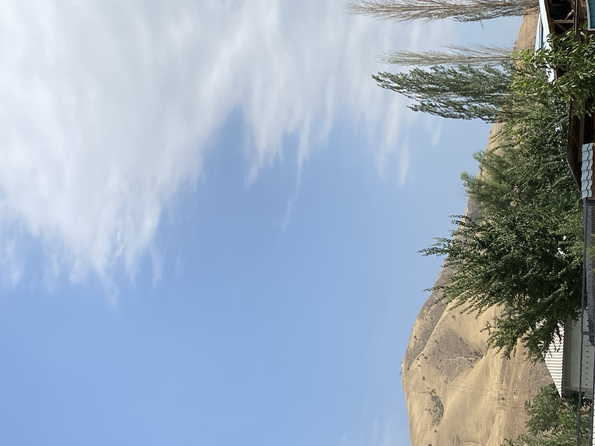 The image depicts a sunny day with a bright blue sky, partially obscured by white clouds on the right side. In the foreground, there is a line of green trees and foliage, with some buildings visible beneath them, featuring corrugated metal roofs. To the left and center of the image, a dry, golden-brown hillside dominates the landscape. On the crest of this hill, a small cluster of white structures, possibly buildings or antennas, are visible. The overall scene suggests a rural or semi-rural environment, potentially on the outskirts of Kara-Kulja, Kyrgyzstan, given the distinctive arid mountain landscape.