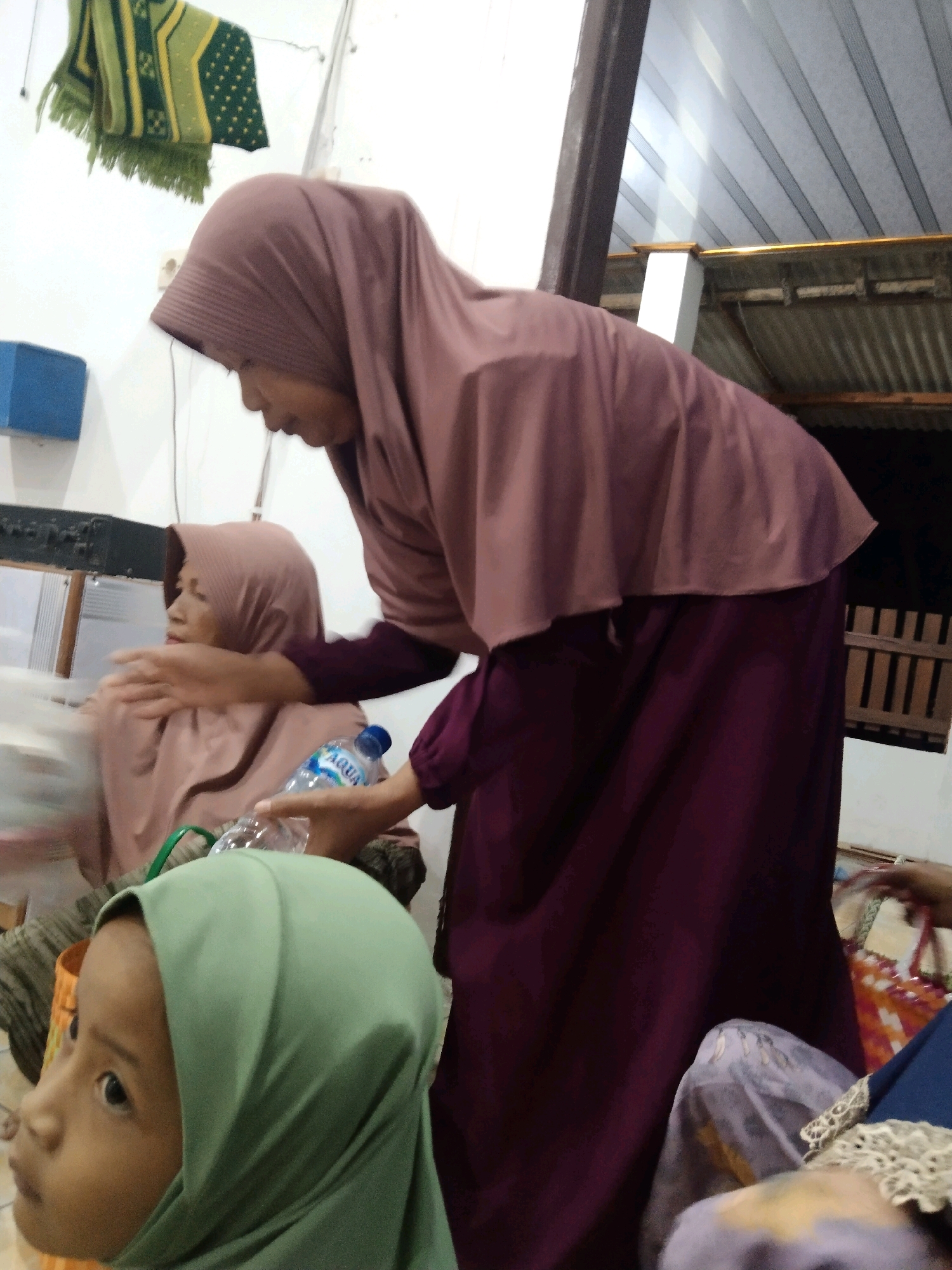 In Buntalan, Indonesia, an indoor scene depicts three individuals, two women and a young child. The woman in the foreground, wearing a mauve hijab and a dark purple dress, is leaning forward and extending her right hand towards another woman. This second woman, seated and also wearing a mauve hijab and a light pink top, is looking to her left. In front of her, a plastic water bottle with a blue cap is visible. To the left of the frame, a young child wearing a light green hijab is partially visible, facing left. In the background, a green and yellow patterned rug hangs on the wall, and corrugated metal roofing is apparent.