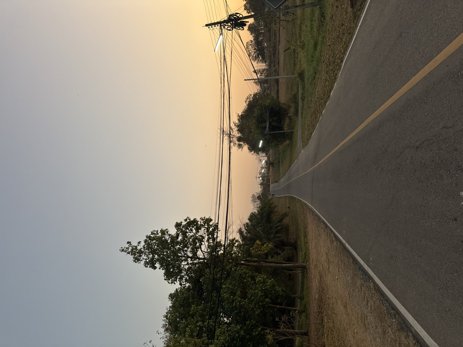 The image depicts an asphalt road ascending uphill and curving slightly left, viewed during early morning or late afternoon in Ban Huai Sai, Thailand. The sky exhibits a gradient from light blue overhead to warm orange and yellow at the horizon. The road features a solid white line on its left edge and a solid yellow center line.
To the left of the road, a strip of dry, reddish-brown earth transitions into dense vegetation, including tall, leafy trees and several palm trees. Overhead, multiple black utility wires stretch horizontally, supported by a wooden utility pole visible on the left.
To the right of the road, the ground is covered in dry grass and sparse green vegetation, bordering what appears to be an open field or agricultural land in the distance. Further along the road on the right, additional utility poles are visible, one featuring an illuminated streetlamp.
In the far distance, indistinct dark figures, possibly two individuals, are visible ascending the road towards the crest of the hill. No immediate interactions or specific actions are discernible among the subjects.