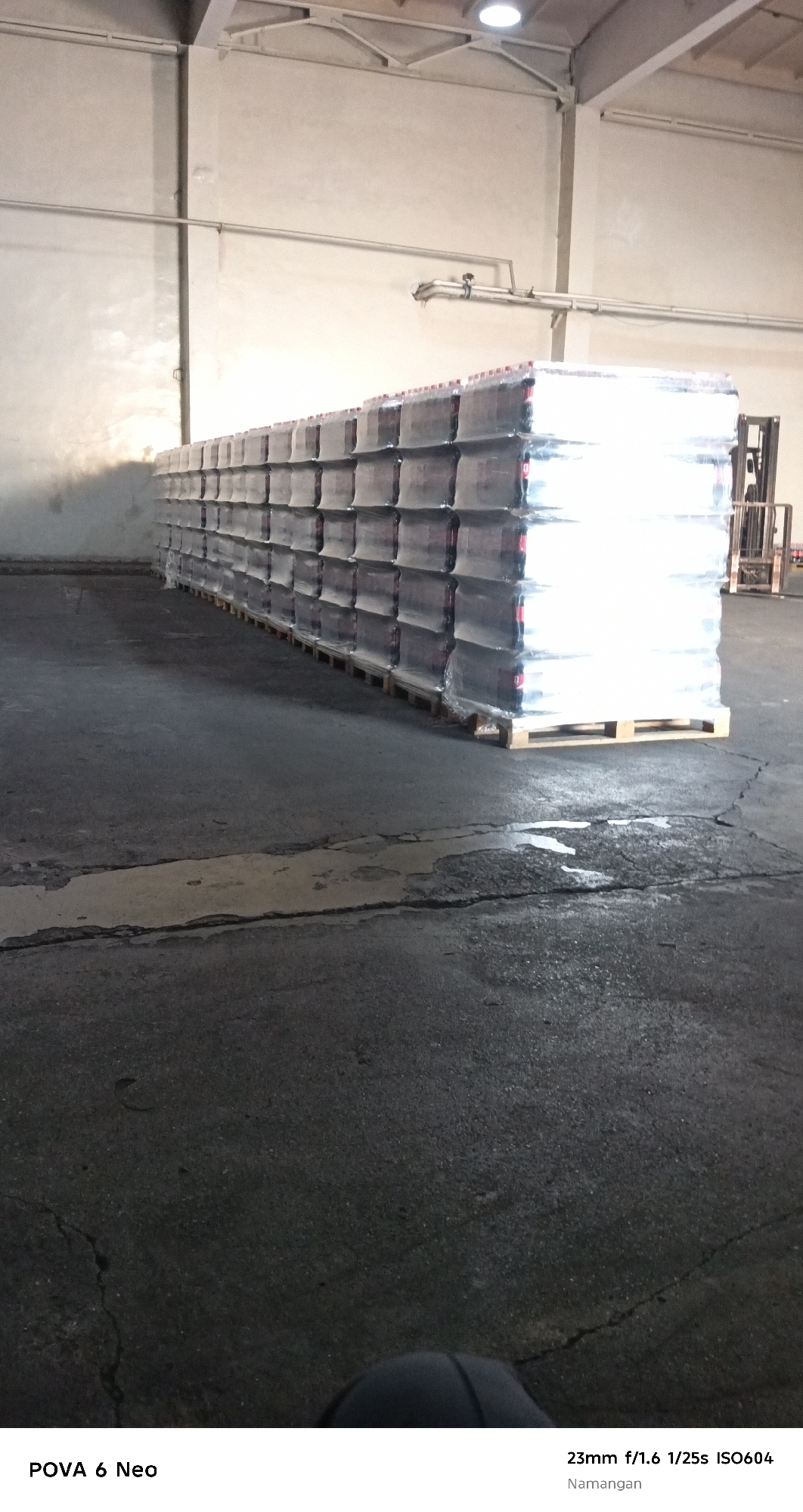 The image depicts a storage area within a warehouse in Namangan City, Uzbekistan. Rows of pallets, each stacked with multiple cases of bottled beverages, are arranged on the concrete floor. The bottles appear to be wrapped in clear plastic. A forklift is partially visible to the right of the stacked pallets. The environment is industrial, with concrete flooring and plain, light-colored walls and ceiling. The lighting suggests artificial illumination within the warehouse. No individuals are visible in the image.
