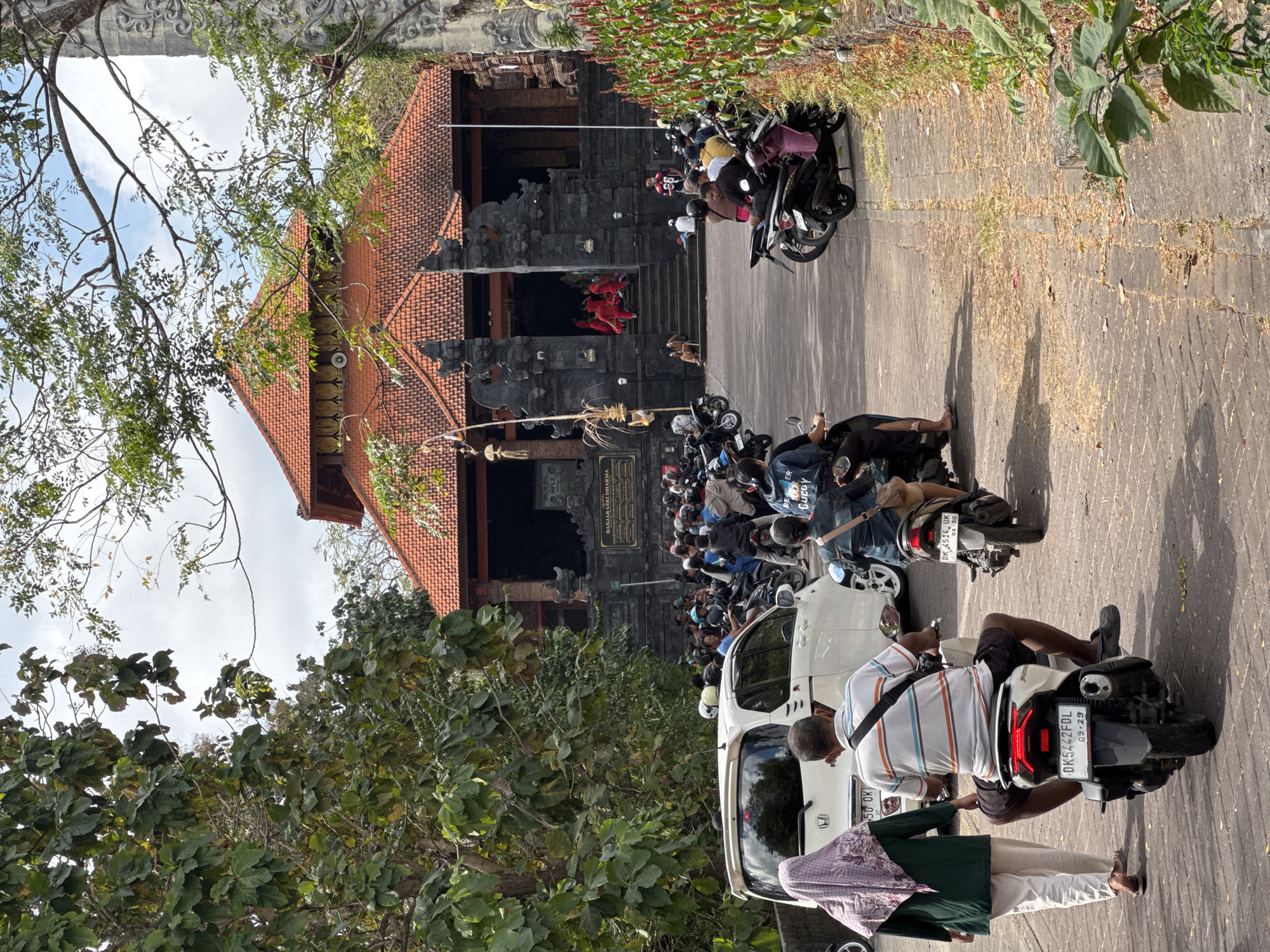 This image captures a lively scene in front of a temple in Kuta Selatan, Indonesia. A crowd of people are gathered, many on motorcycles, in a paved area leading up to a temple with a red tiled roof and dark stone architecture. Several individuals in red attire are visible on the steps of the temple, suggesting a possible ceremony or cultural event. The environment is characterized by lush green trees and vegetation lining the sides of the path, with a sunny sky overhead. The presence of numerous motorcycles indicates this is a popular location, likely a tourist destination or a significant local landmark.
