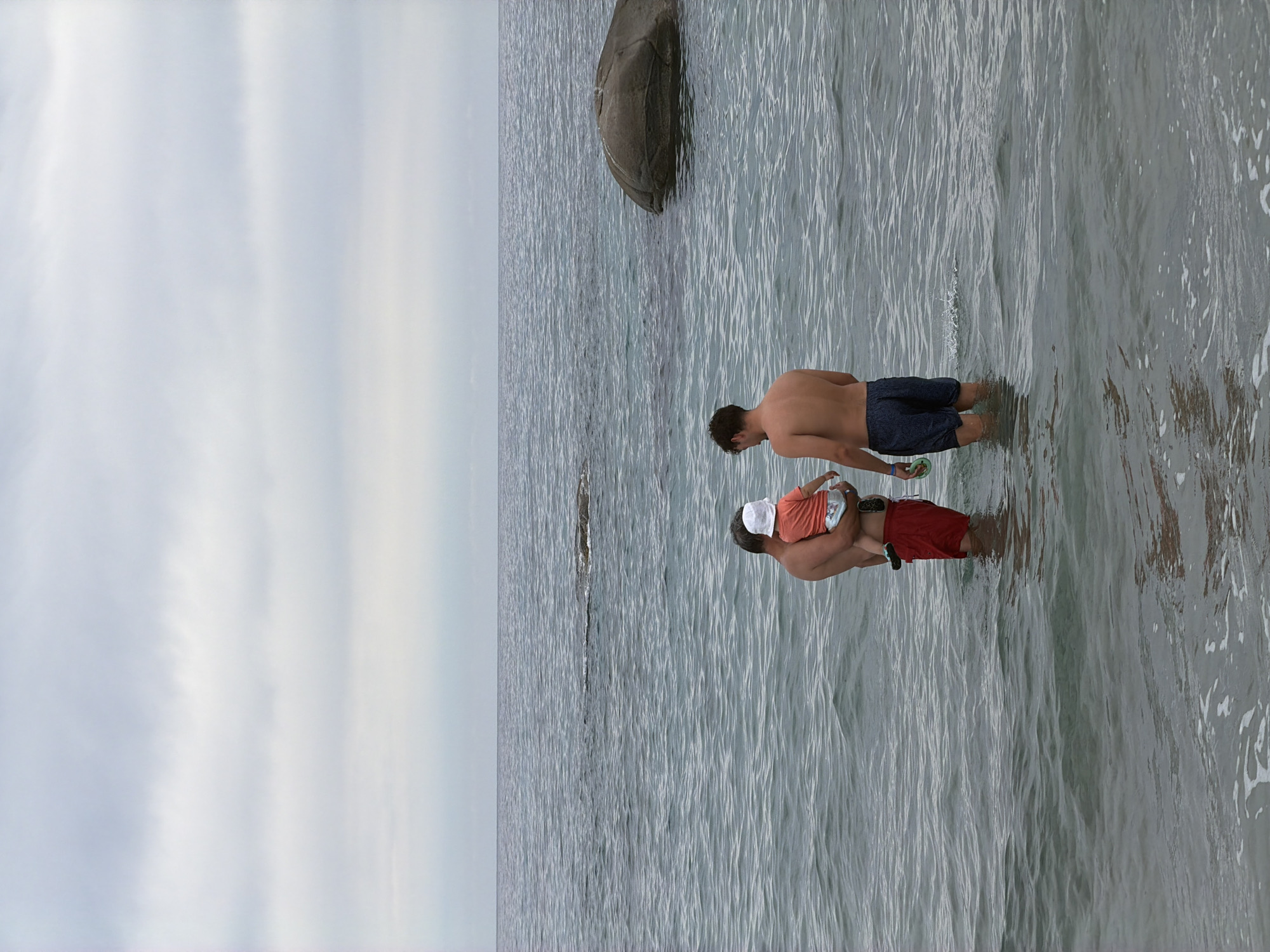 Two men and a child stand in the calm ocean water off the coast of Tortolì, Italy. The man on the left, facing away from the camera, holds a young child dressed in an orange shirt and white hat. He wears red swim trunks. The man on the right, also facing away, wears dark blue swim trunks. The sky above is overcast with layers of grey clouds. The sea is a muted blue-grey, with gentle waves. In the distance, two large rocks are visible protruding from the water, one on the left and a much larger one on the right. The overall scene depicts a relaxed moment at the beach under a cloudy sky.