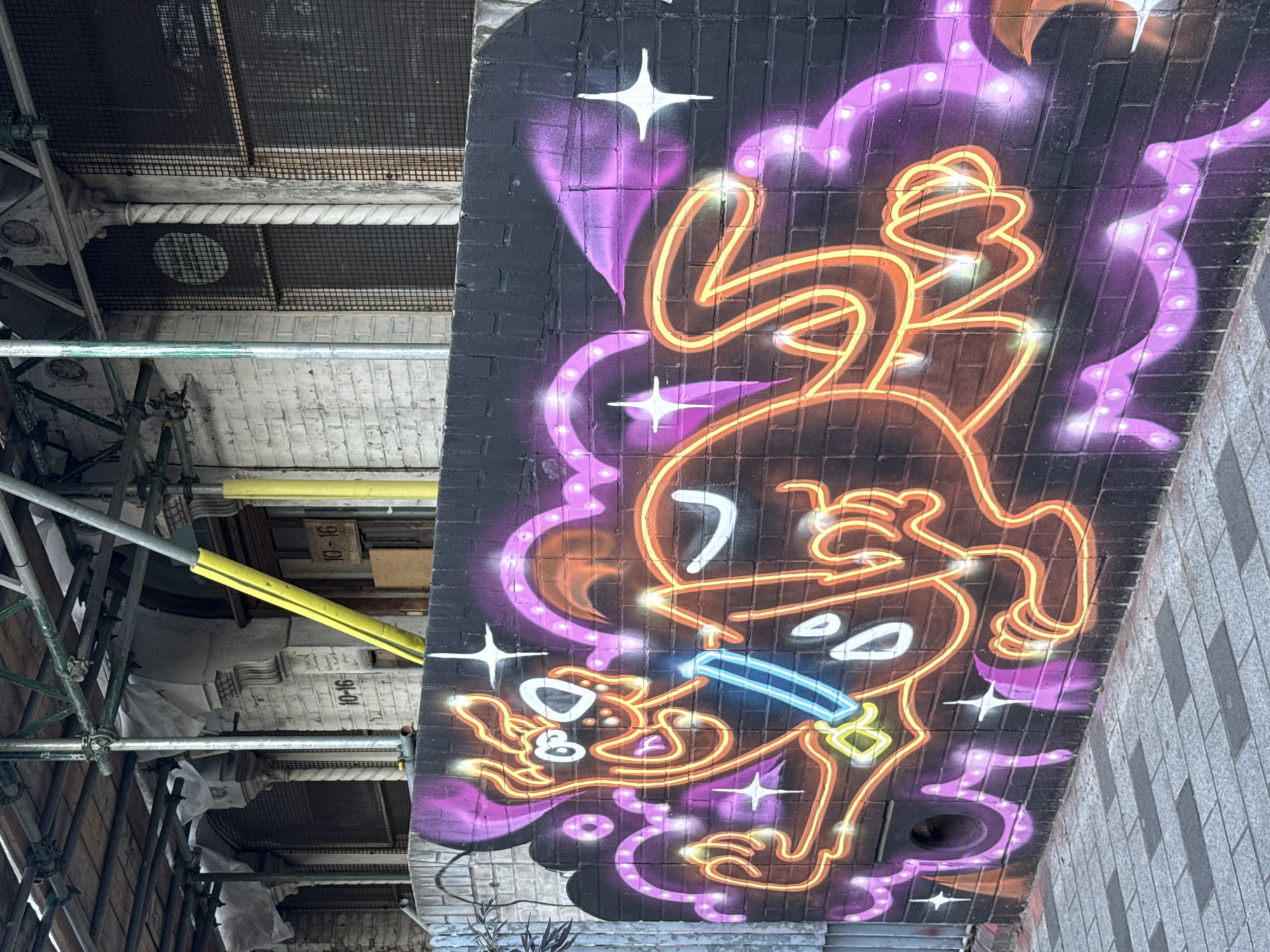 A low-angle shot captures a vibrant graffiti artwork painted on the dark brick underside of a bridge or underpass, contrasting sharply with the surrounding urban infrastructure. The central subject of the graffiti is a stylized, cartoon-like character, possibly a dog or rabbit, rendered with bright orange and yellow "neon" outlines that give it a glowing effect. This character is depicted in a dynamic, playful pose, seemingly jumping or dancing, with white oval eyes and holding a glowing blue stick or object in its mouth. Surrounding the character are purple cloud-like shapes with white dots and sharp white starbursts, further enhancing the luminous, energetic aesthetic.
To the left of the artwork, an intricate network of metal scaffolding with yellow protective padding is prominent, suggesting ongoing construction or maintenance. Through the scaffolding, various structural details are visible, including textured white walls, dark mesh grates, and a decorative twisted column. A gray tiled wall is present on the far right of the image.
The scene conveys an urban environment, indicative of a city like London, United Kingdom. The vivid, almost fluorescent colors of the graffiti provide a striking artistic focal point against the muted, functional tones of the brickwork, metal, and concrete. Visible text includes "10-16" on several wooden planks within the scaffolding and a faint "14" on a circular white patch on a mesh grate under the bridge. The image appears to be taken during the daytime, with natural light illuminating the detailed artwork and its industrial surroundings.