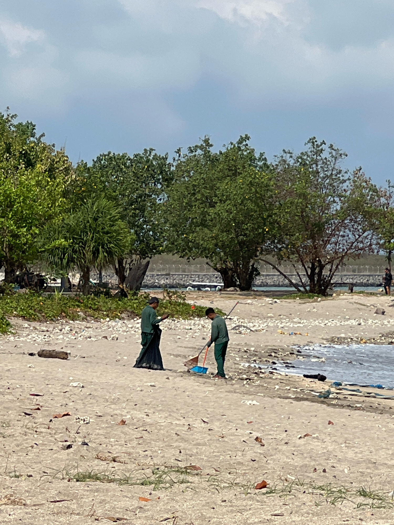 In Kuta, Indonesia, two individuals in green uniforms are cleaning a littered beach. One person is using a broom and dustpan, collecting debris into a blue bucket. The other person stands beside them, holding a black garbage bag. The beach is covered with various trash, including plastic bottles, wrappers, and other waste. In the background, lush green trees line the shore, with a stone wall and the ocean beyond. The sky is overcast with light clouds.